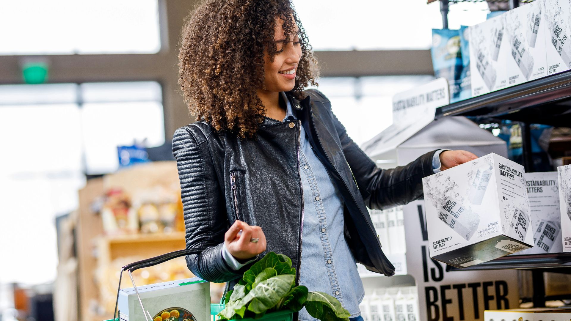 A woman carrying a grocery basket of vegetables picks up a Boxed Water box