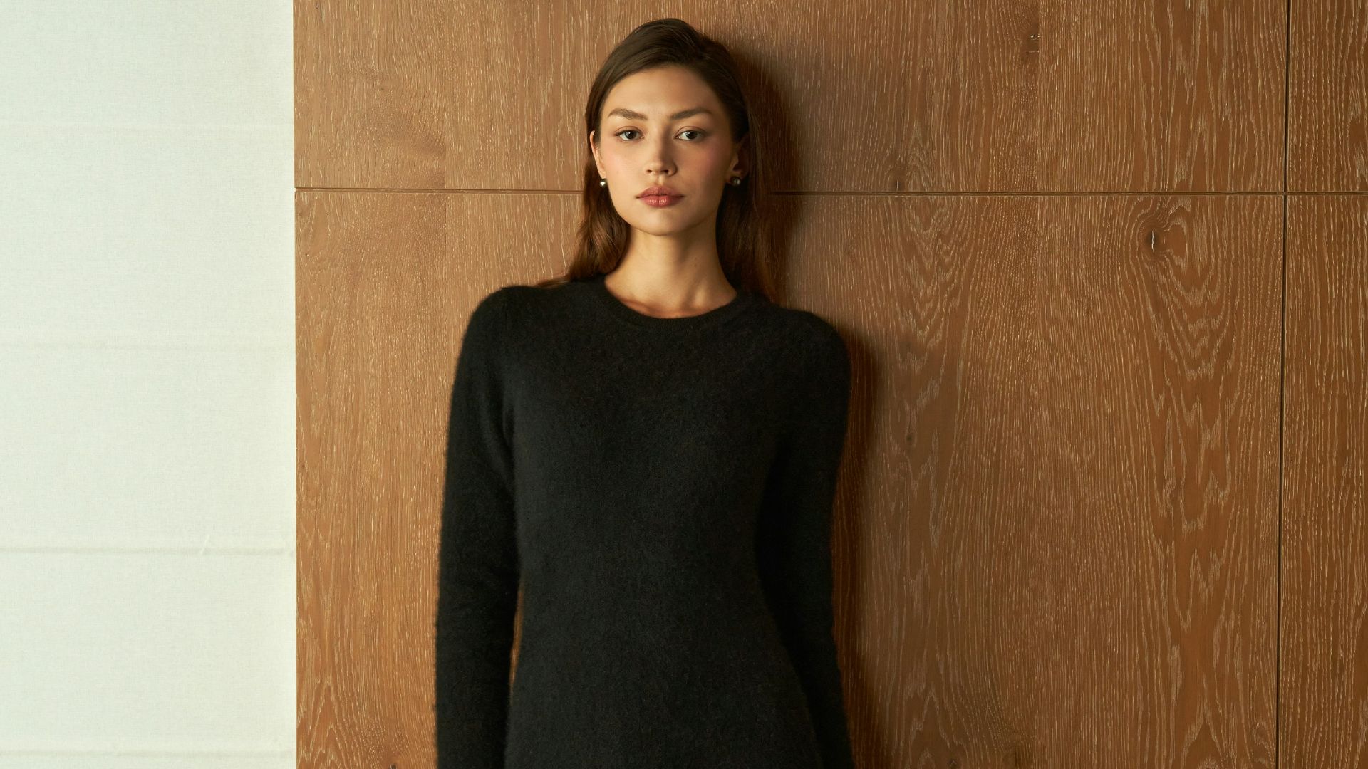 Young woman in a black dress standing against wood paneling.