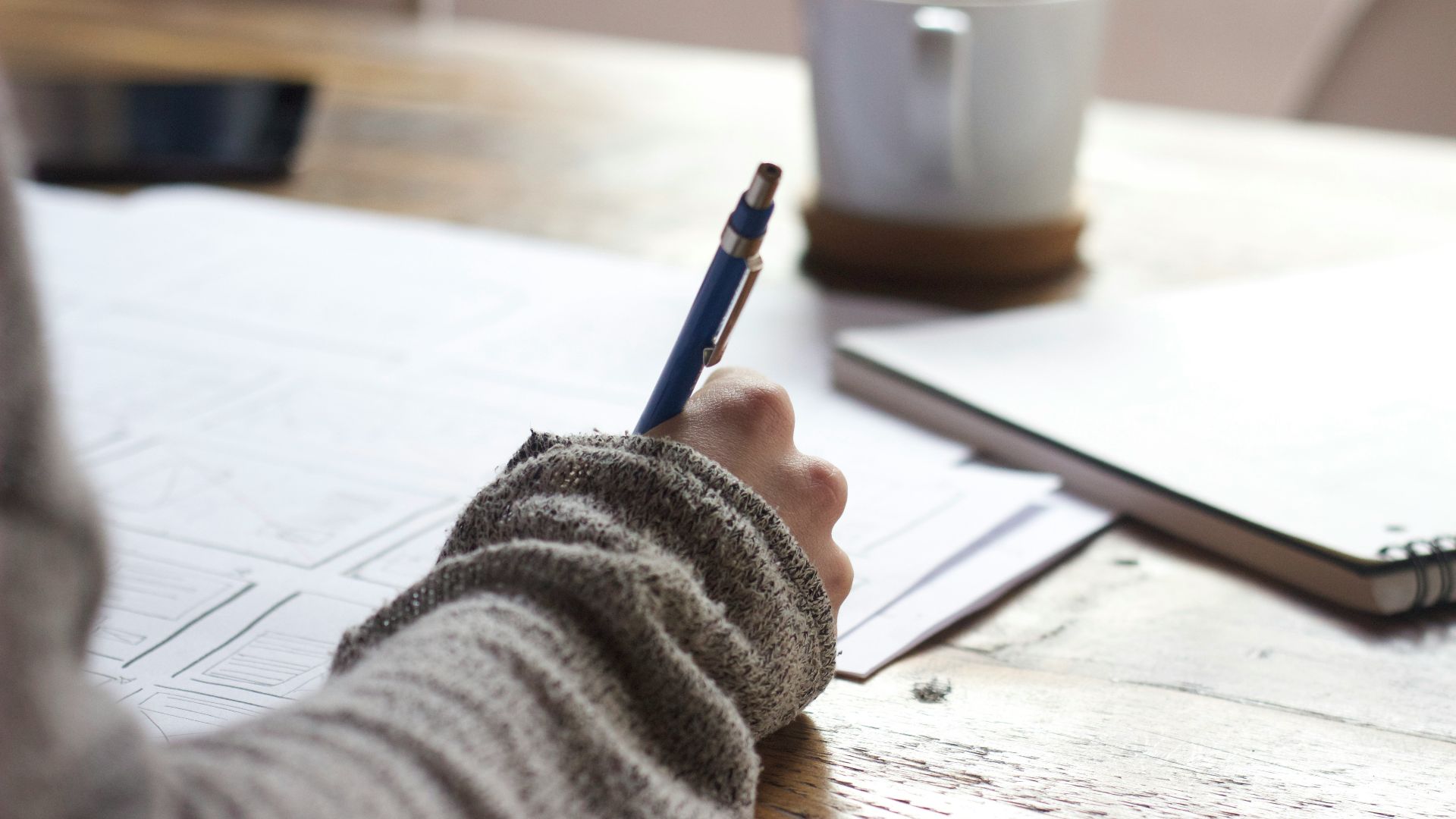 person writing on brown wooden table near white ceramic mug