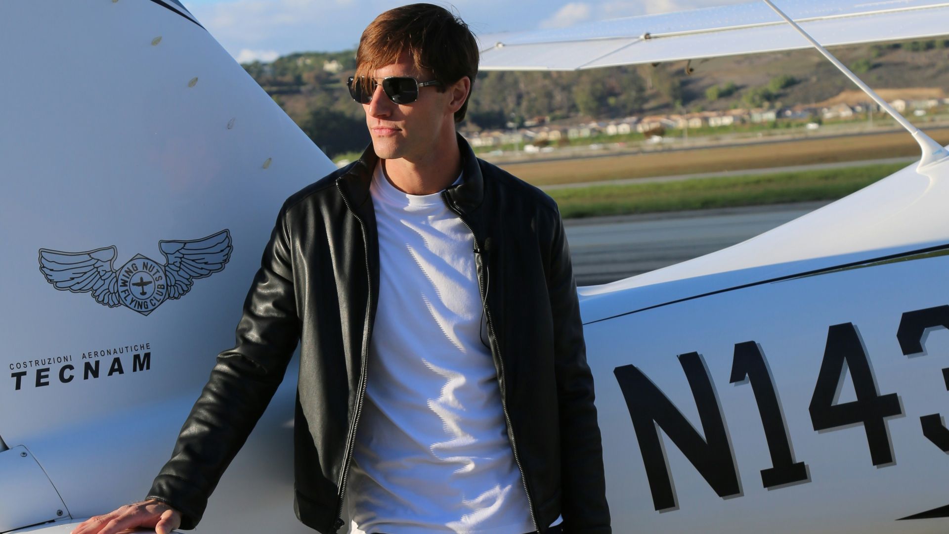 Man in leather jacket poses by a small airplane.