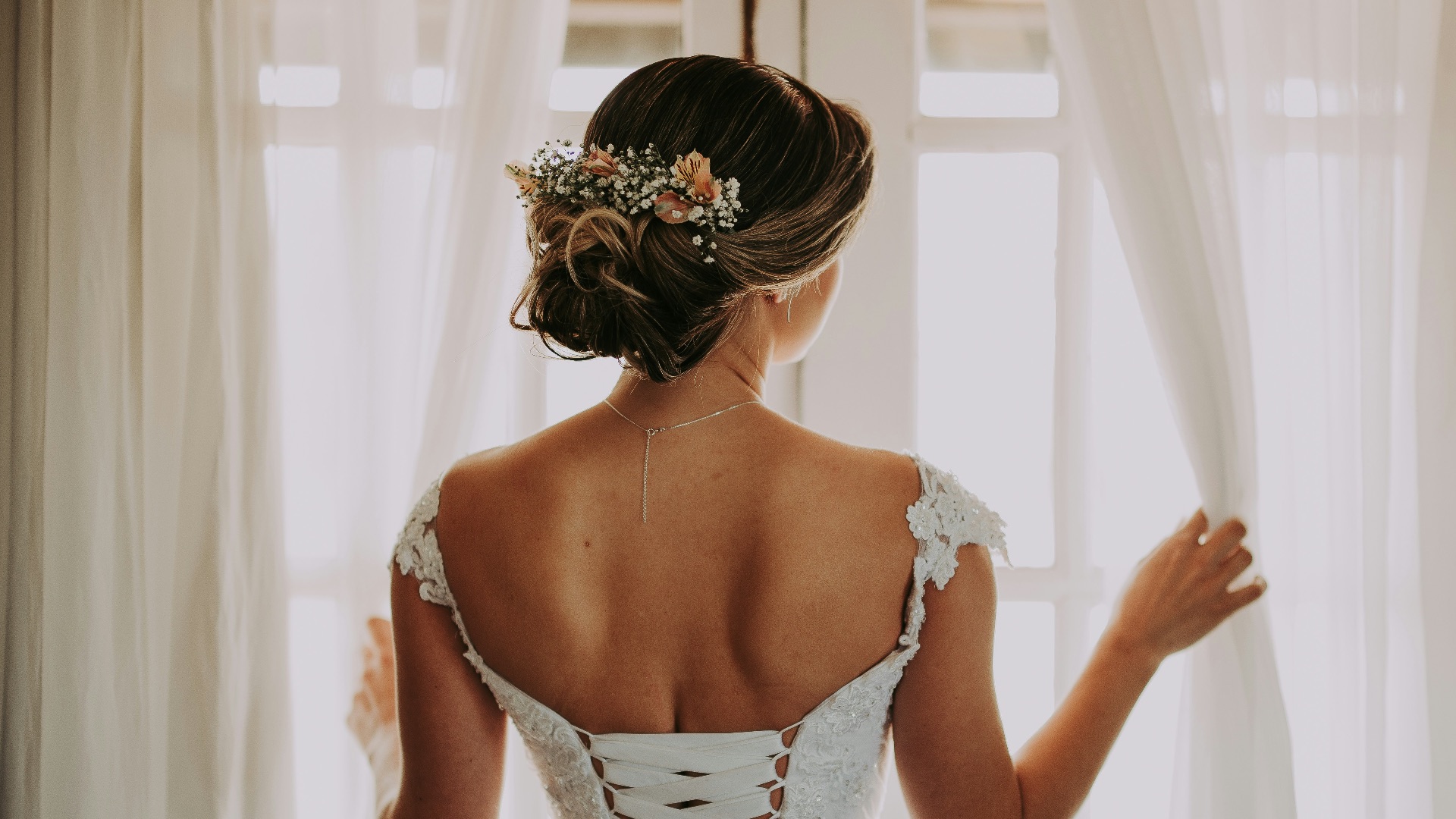 woman in wedding dress standing near window looking outside while holding the curtains
