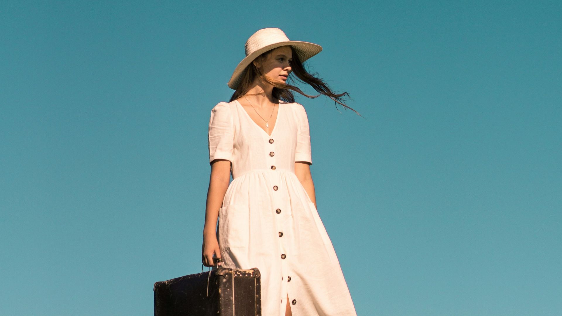 woman standing on green grass holding brown leather suitcase wearing white button-up dress