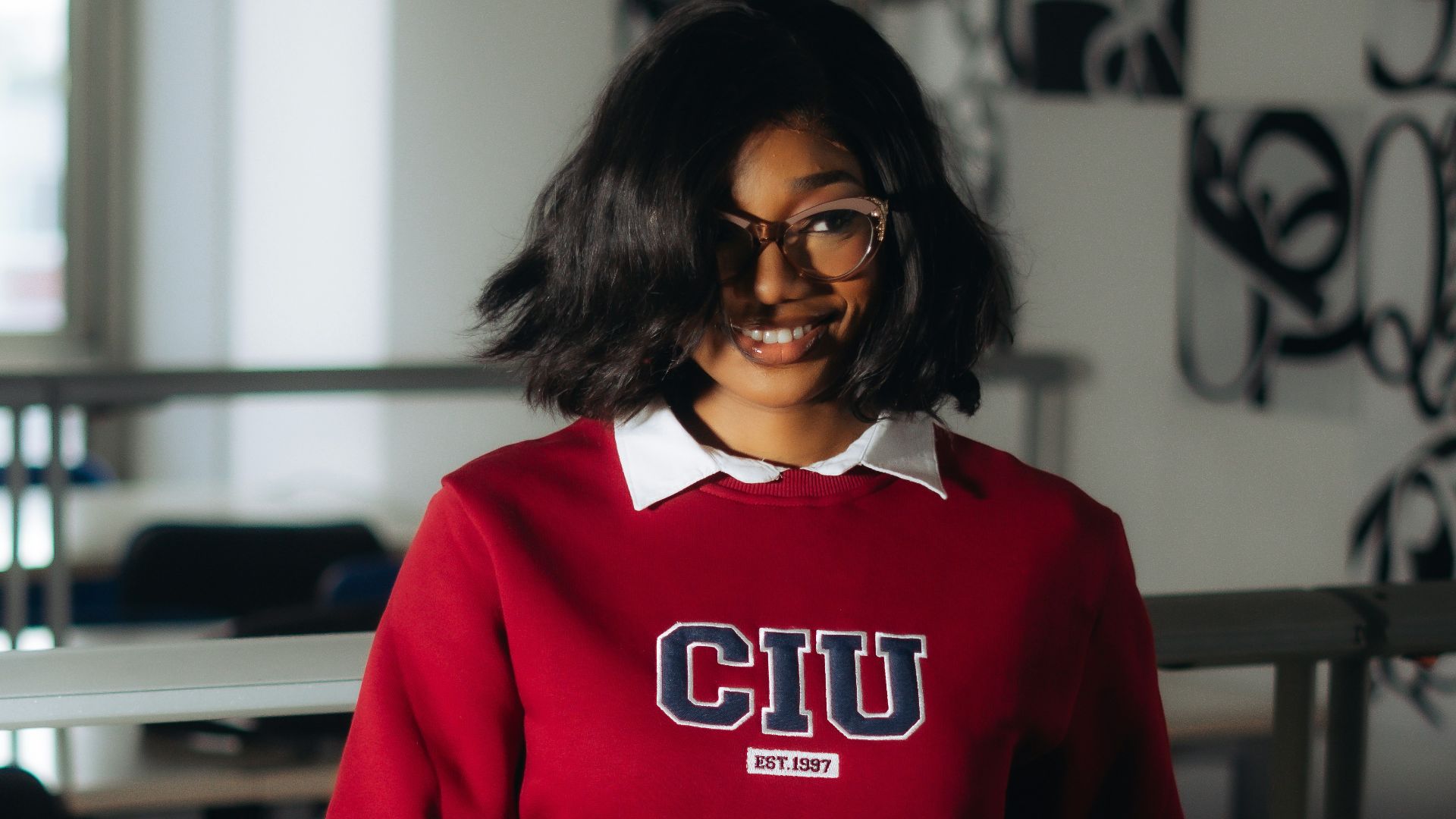 Young woman in glasses and red sweater sits at desk.