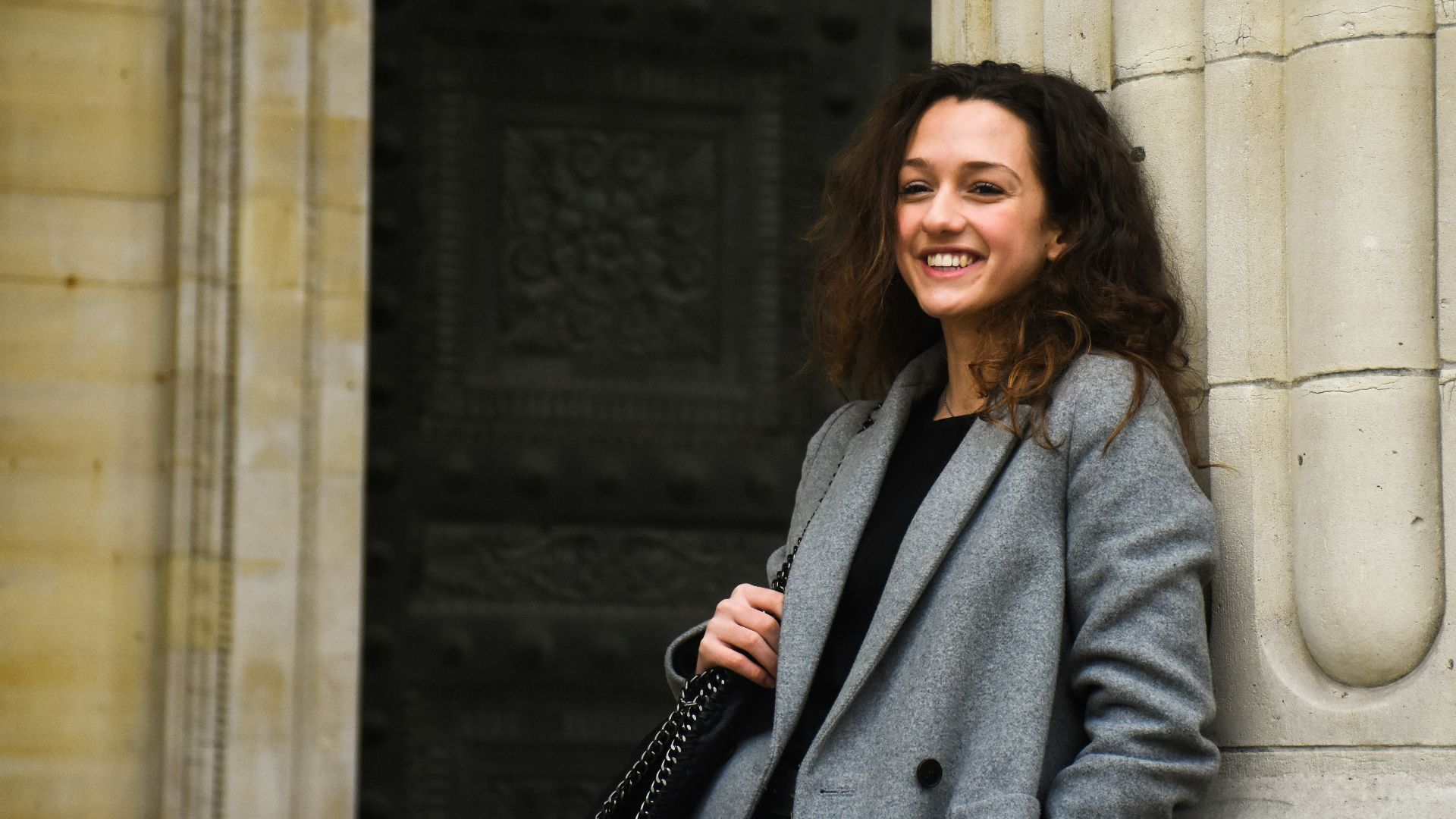 smiling woman holding black shoulder bag leaning on white wall