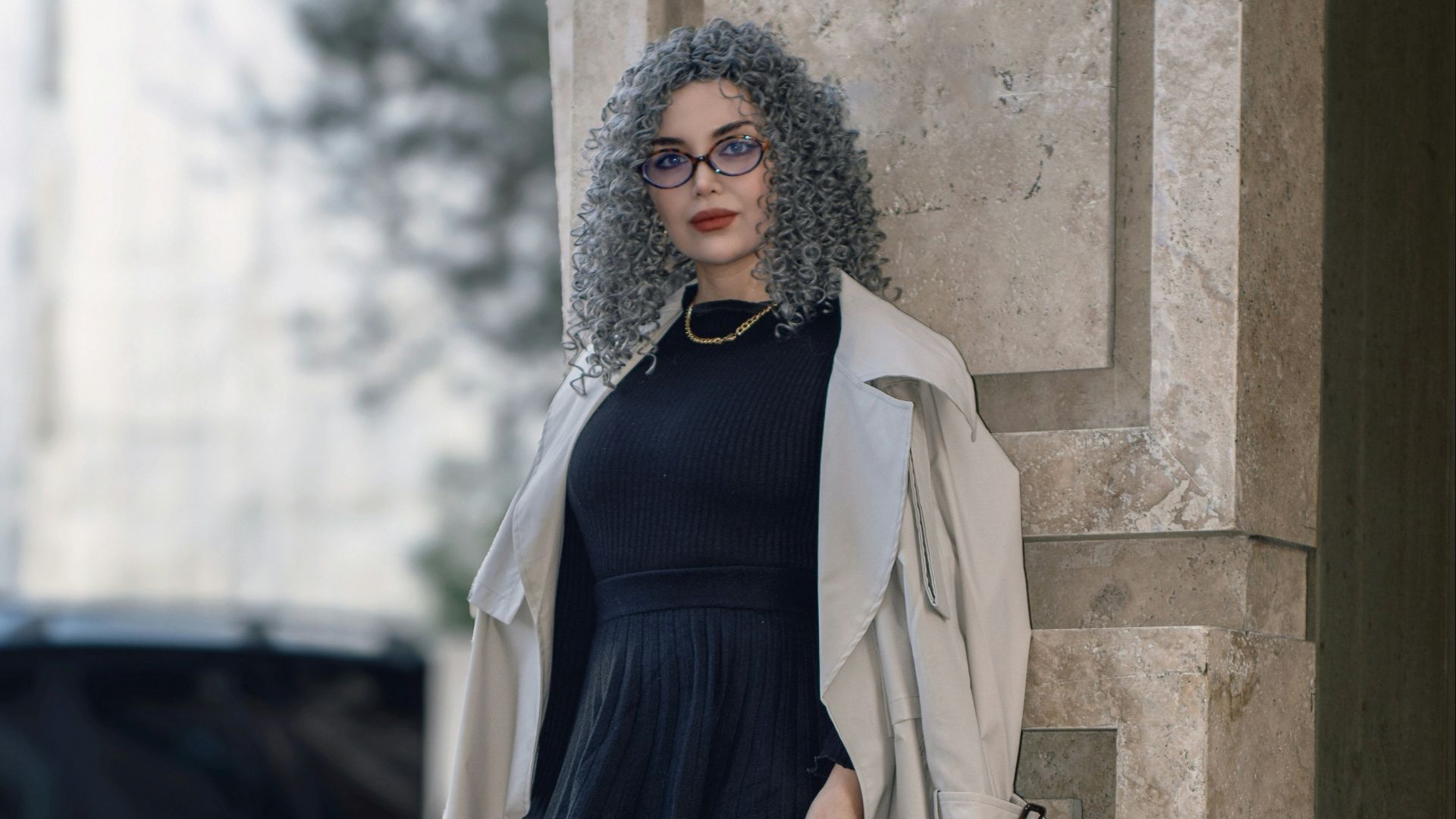 Woman in trench coat and boots leaning on column.