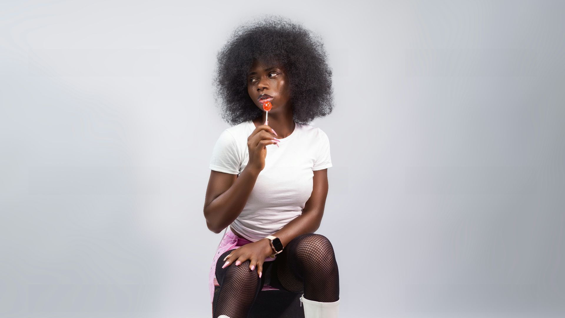 Woman with afro hair wearing white boots, holding lollipop.