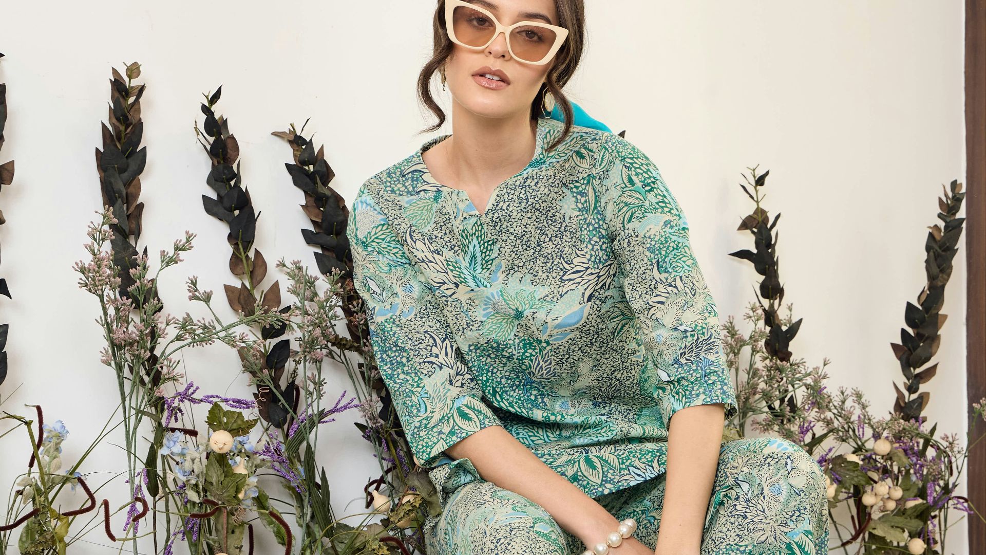 Woman in patterned outfit and sunglasses sits near plants.