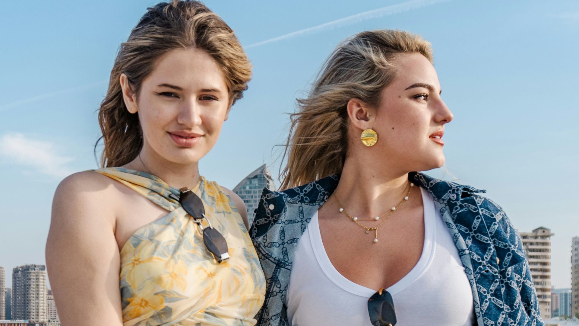 Two young women posing outdoors with cityscape background.