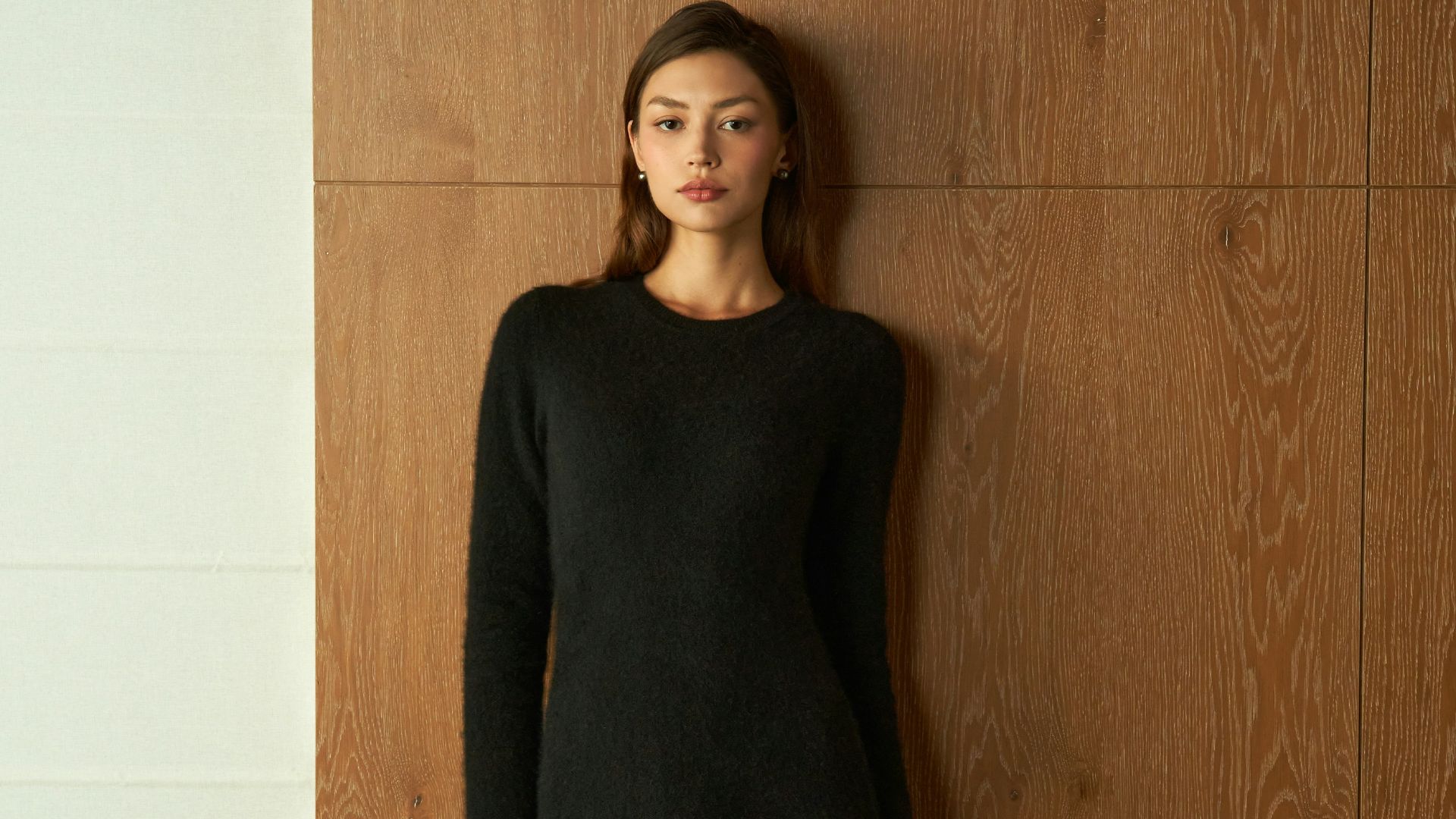 Young woman in a black dress standing against wood paneling.