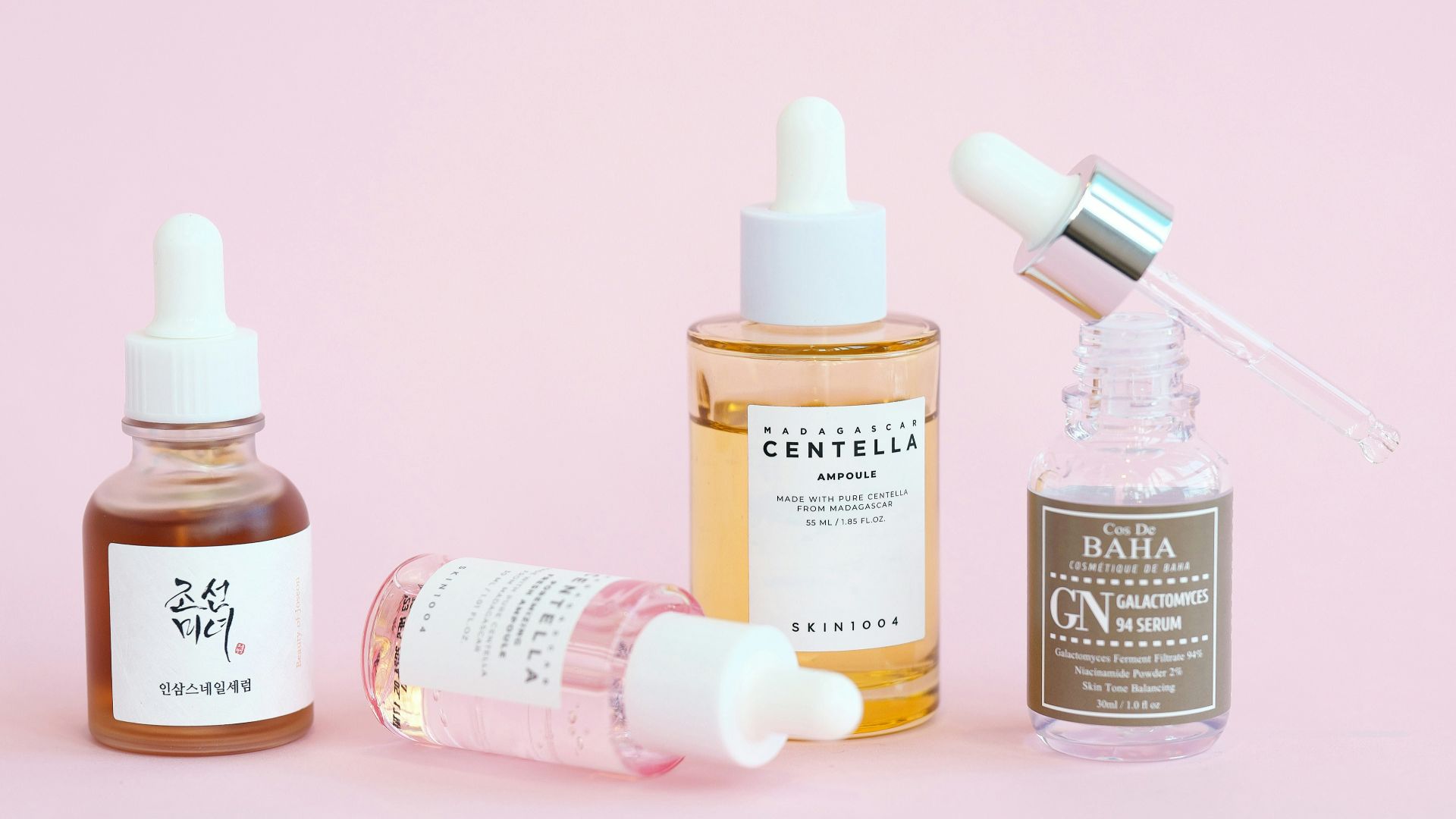 Skincare serums and dropper bottles sit on pink.
