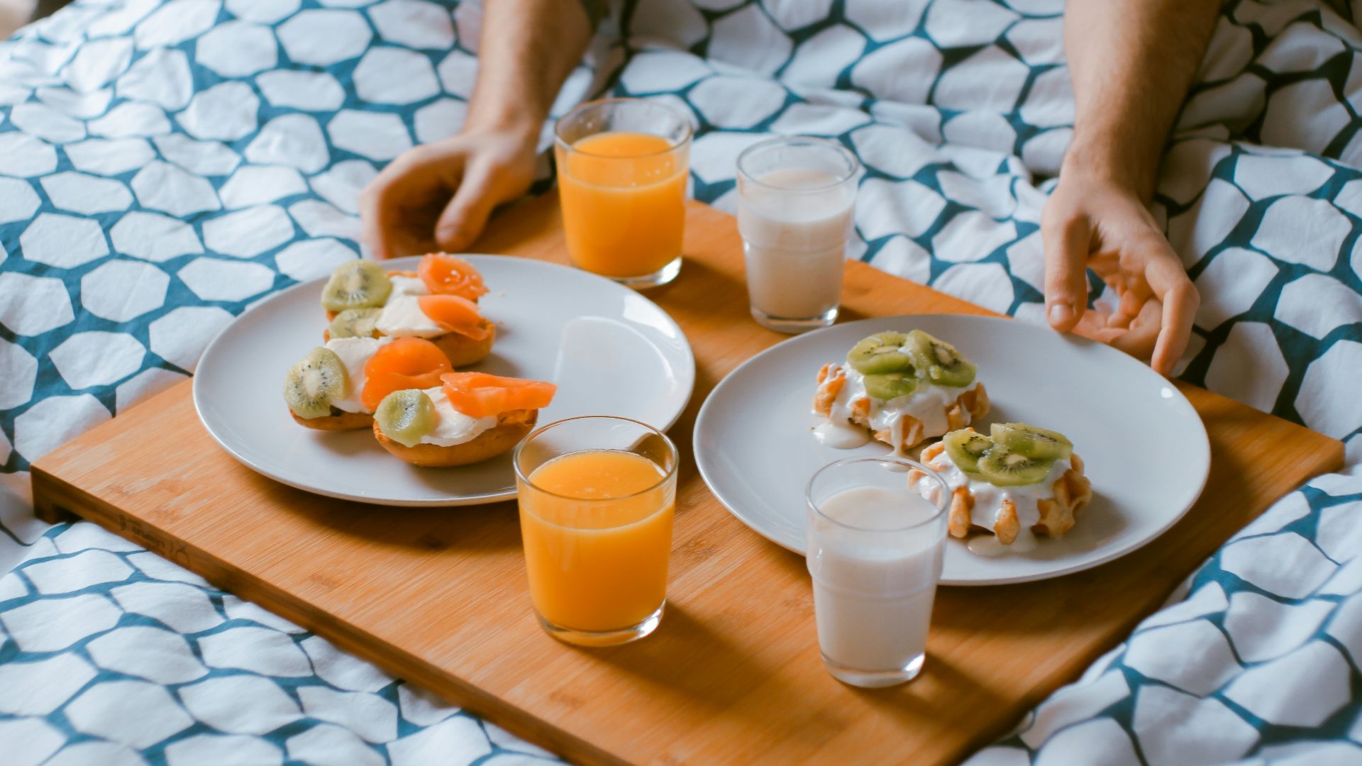 person serving pastries on white ceramic plates with fruit juice glasses on wooden tray on top of bed