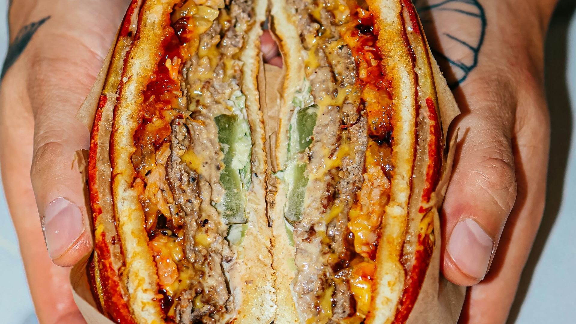 Hands hold a large, sliced burger with pickles.