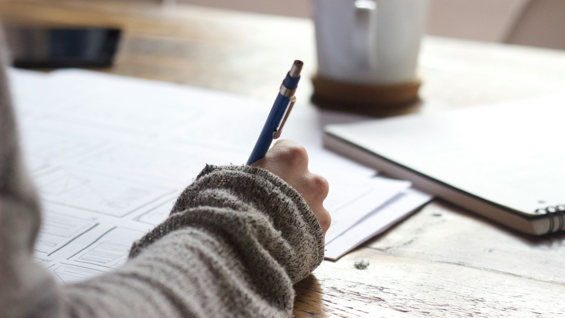 person writing on brown wooden table near white ceramic mug