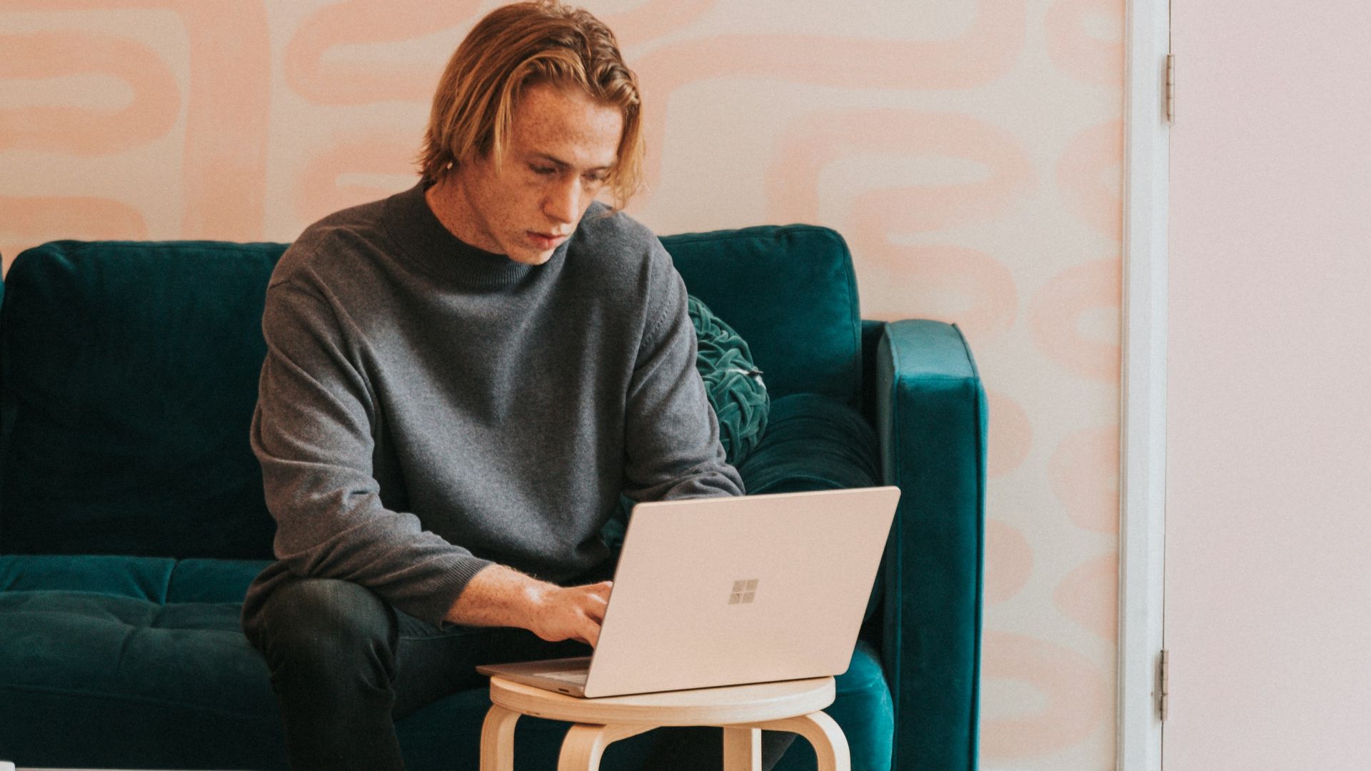 man in gray long sleeve shirt sitting on blue sofa working on a Microsoft Surface laptop