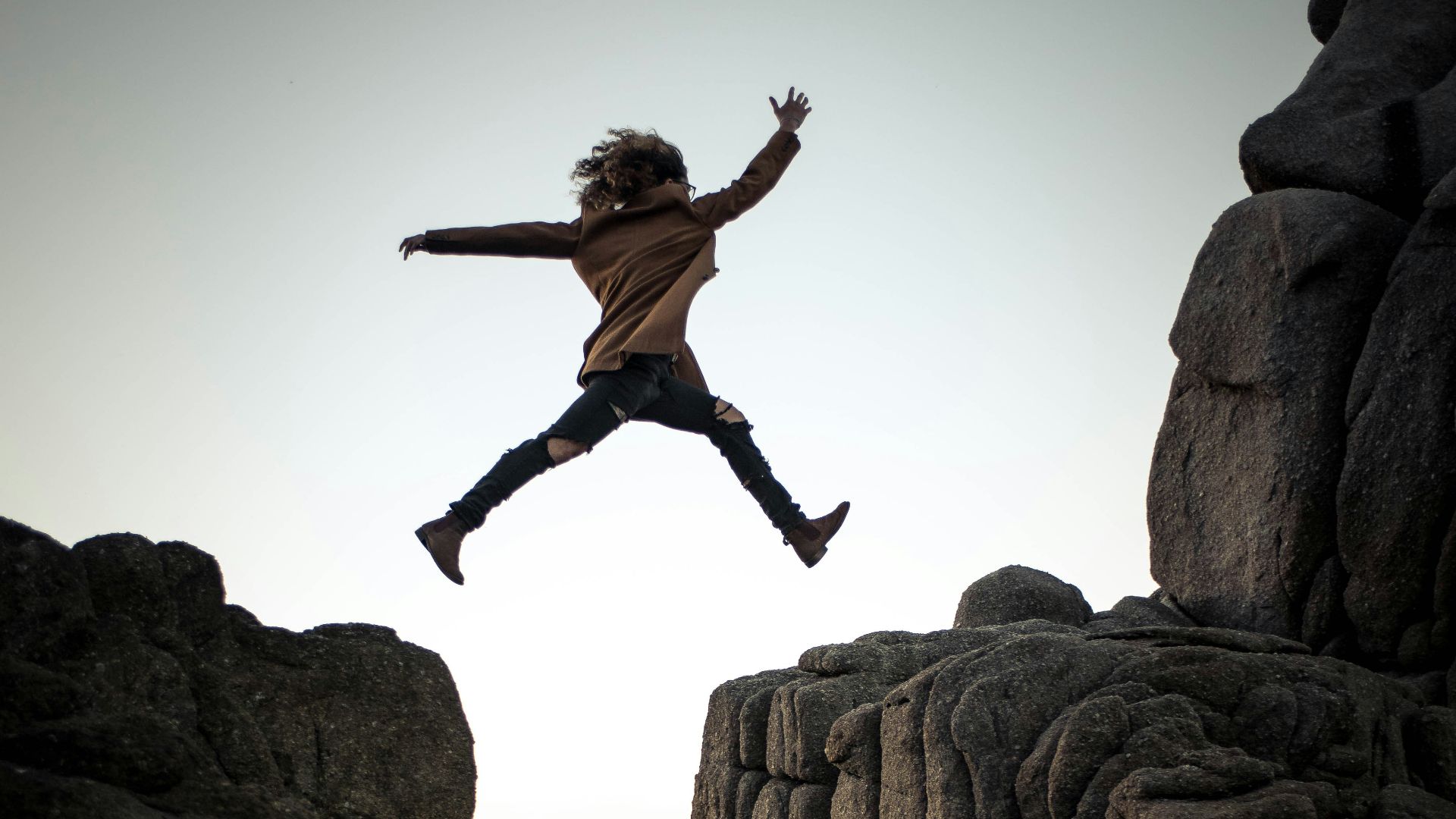 person jumping on big rock under gray and white sky during daytime