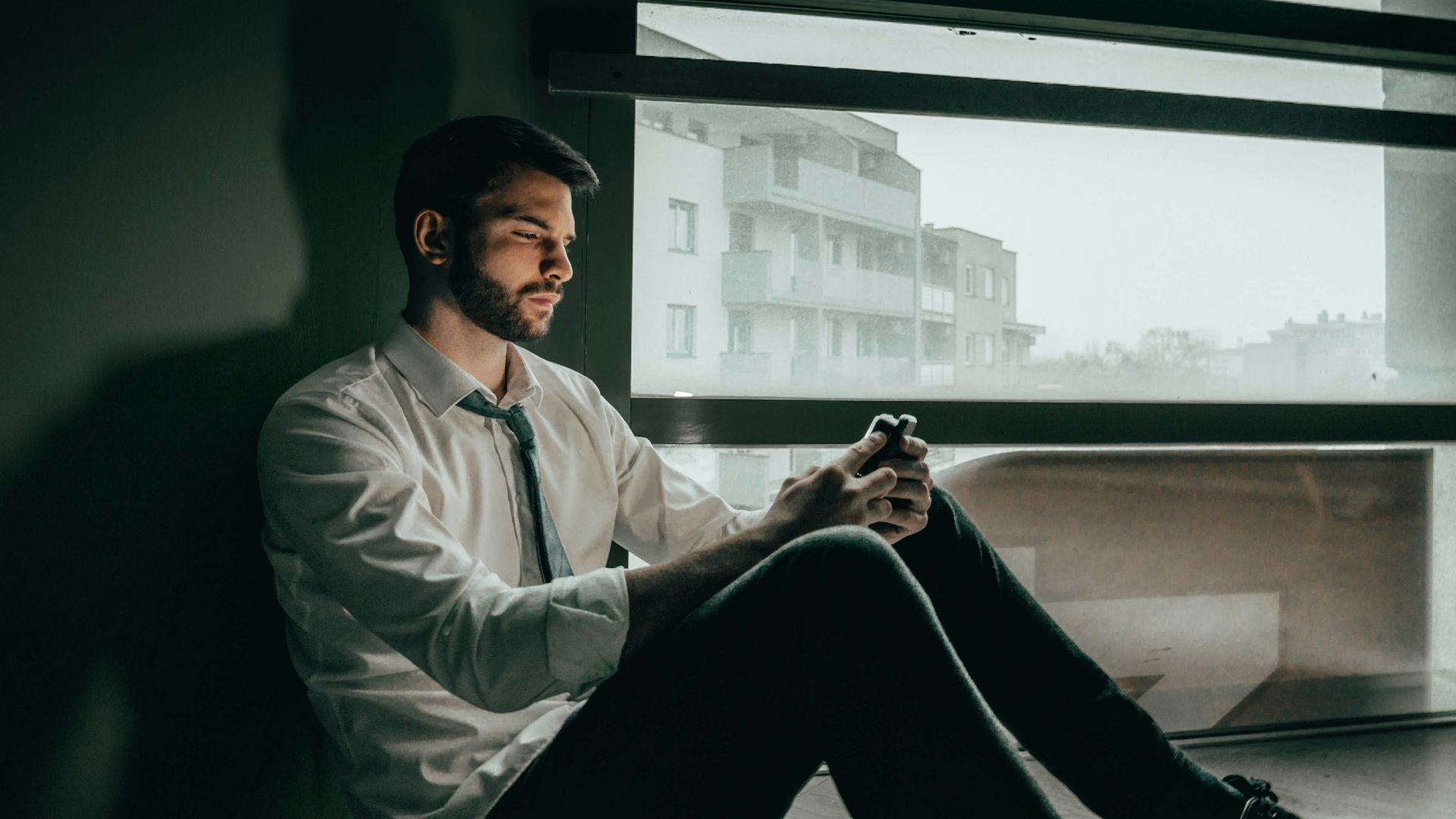 a man sitting on a window sill looking at his cell phone