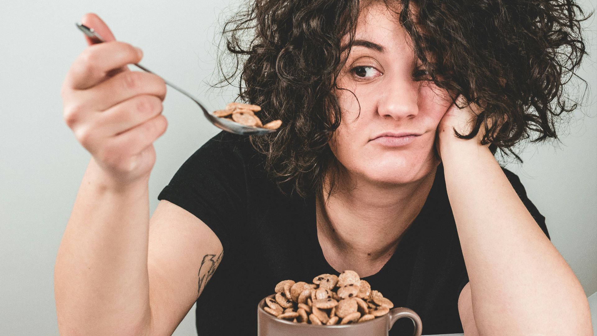 woman with messy hair wearing black crew-neck t-shirt holding spoon with cereals on top