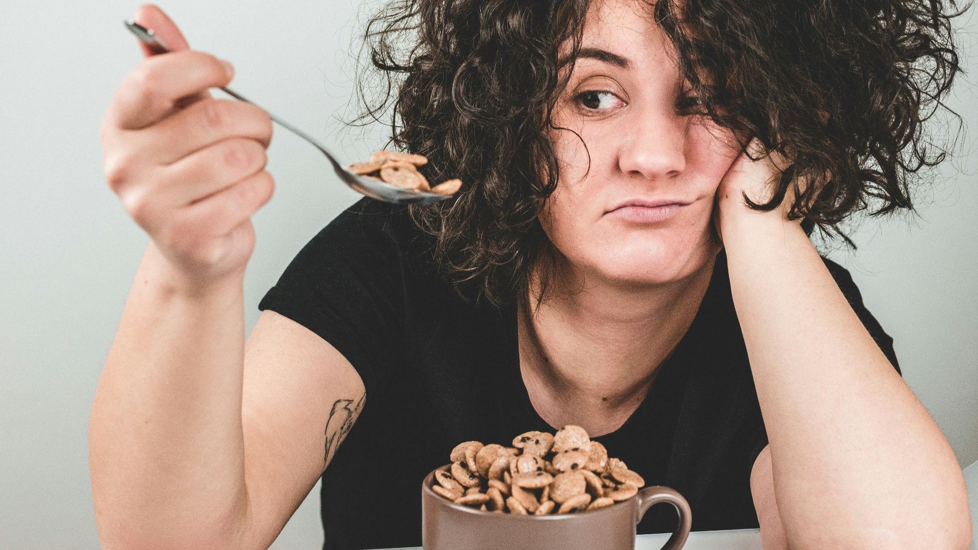 woman with messy hair wearing black crew-neck t-shirt holding spoon with cereals on top