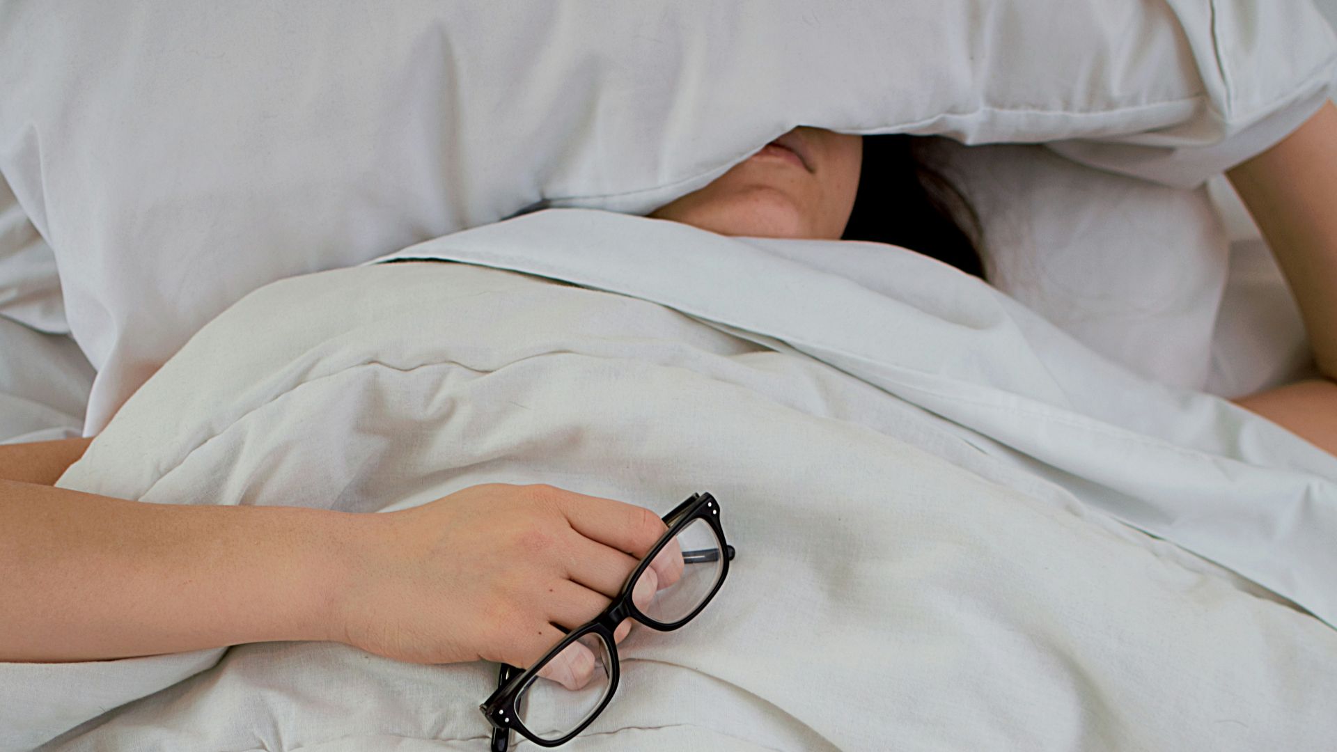person lying on bed while covering face with pillow and holding eyeglasses