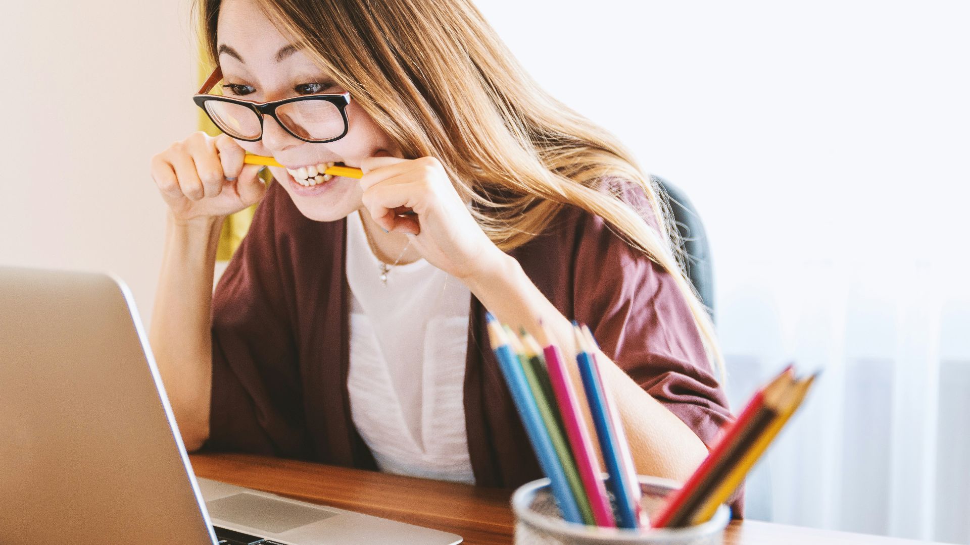 woman biting pencil while sitting on chair in front of computer during daytime