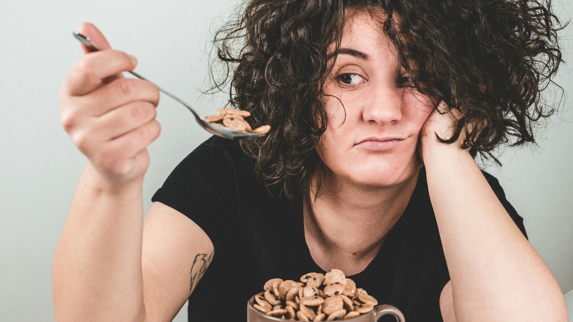 woman with messy hair wearing black crew-neck t-shirt holding spoon with cereals on top