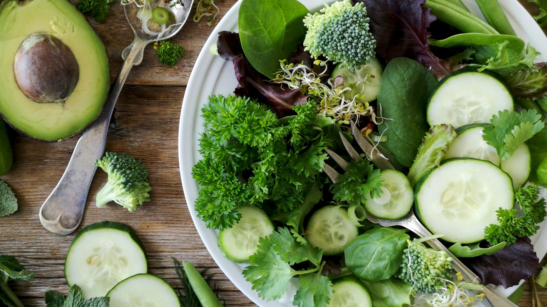 sliced broccoli and cucumber on plate with gray stainless steel fork near green bell pepper, snowpea, and avocado fruit