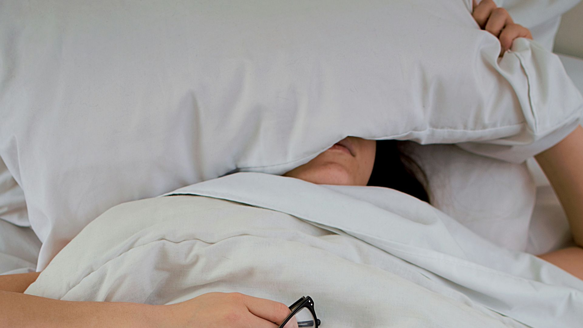 person lying on bed while covering face with pillow and holding eyeglasses