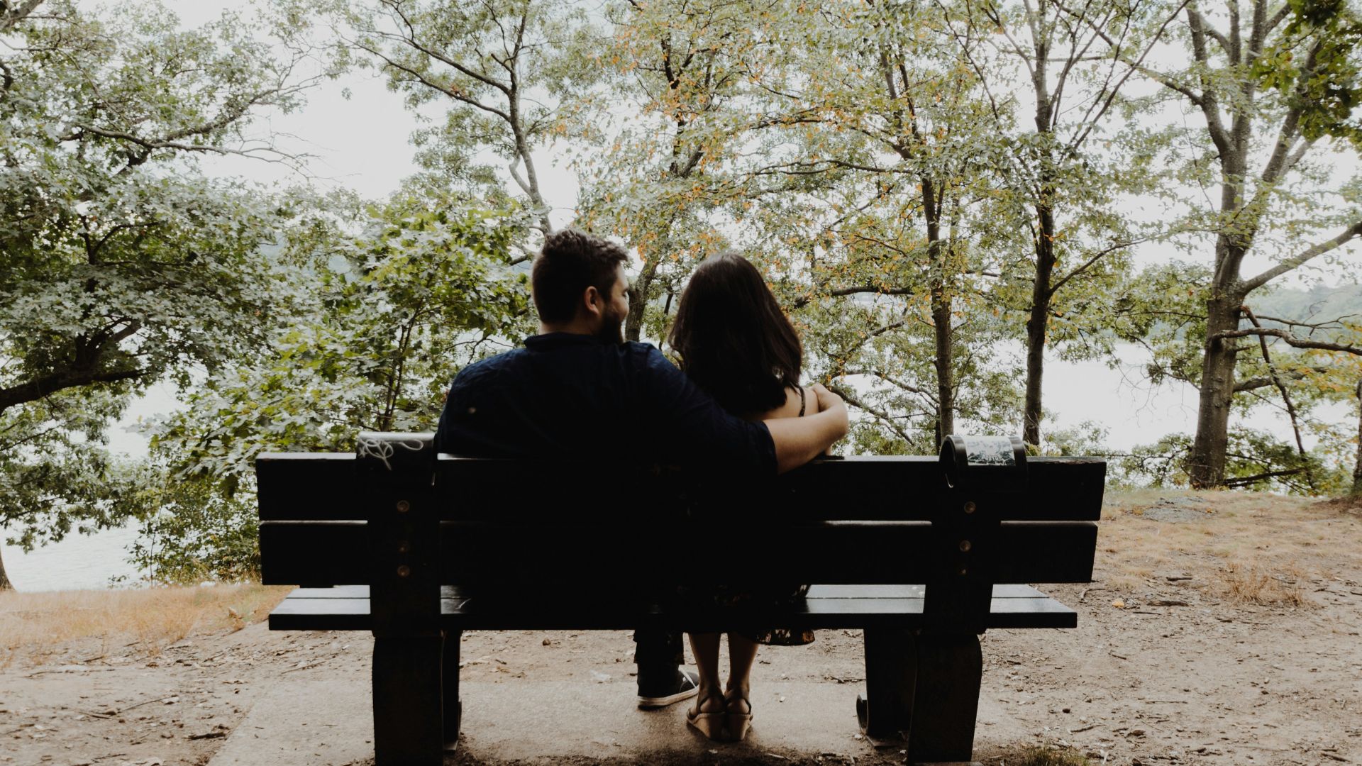 man looking to woman sitting on black wooden bench in front of tall trees during daytime