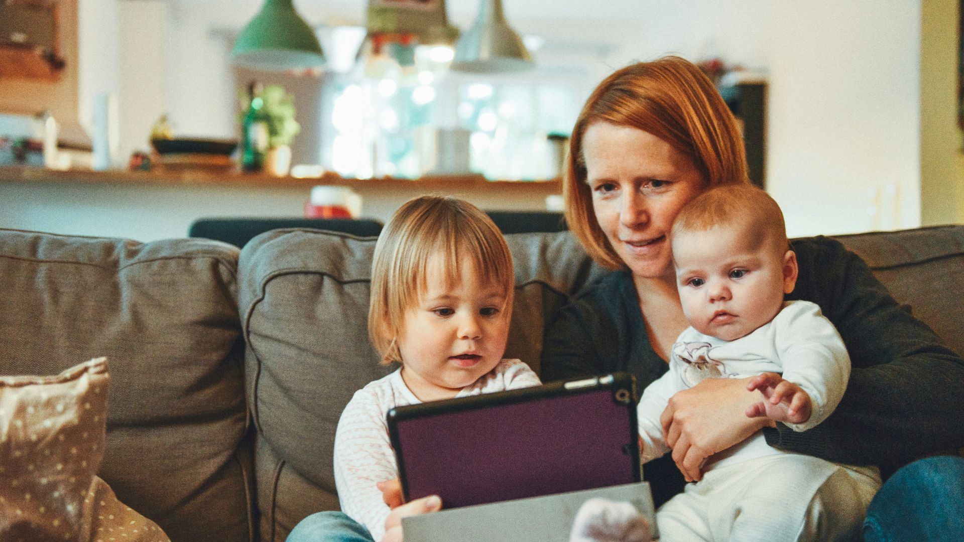 two babies and woman sitting on sofa while holding baby and watching on tablet