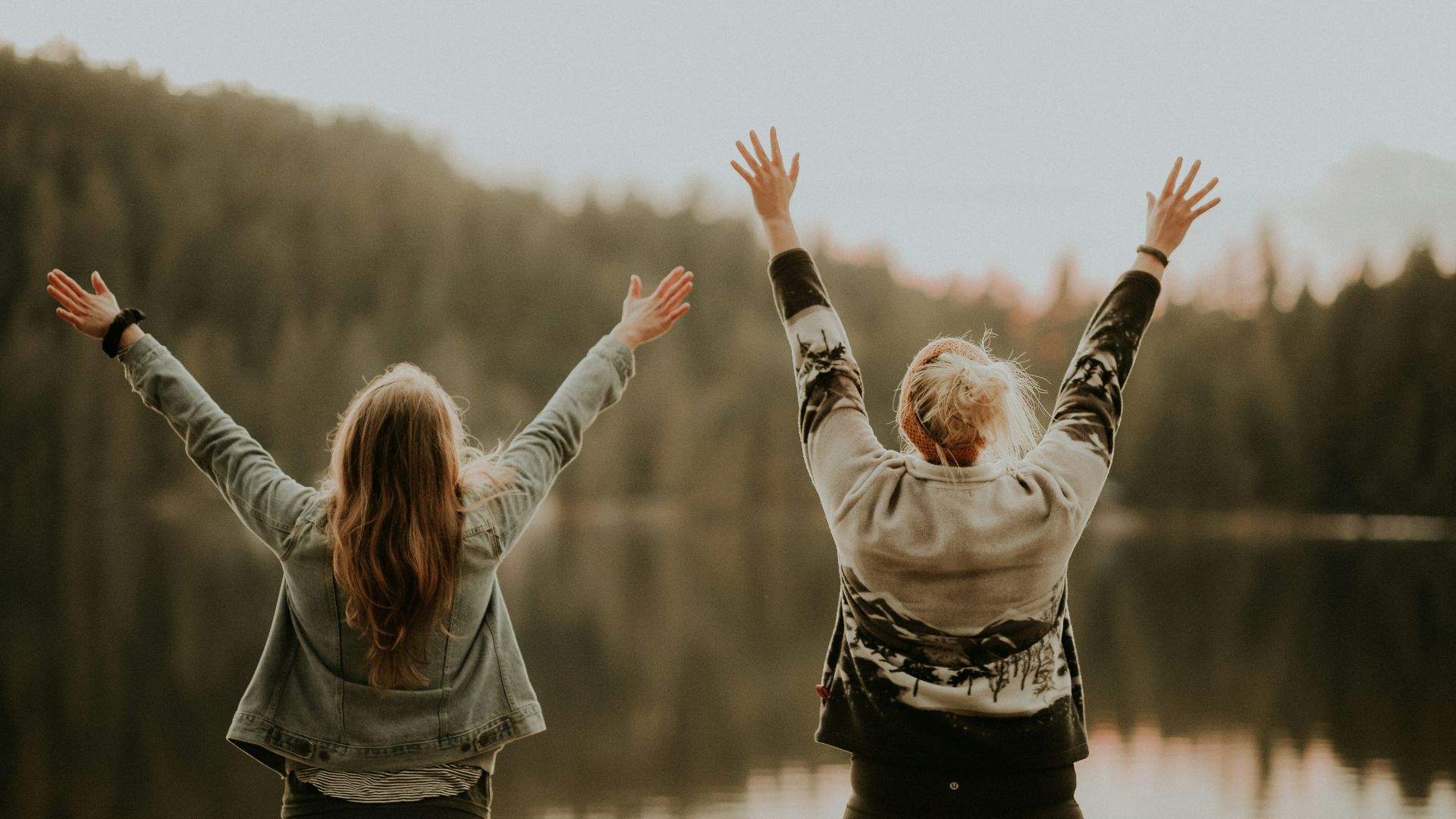 two women hands up standing beside body of water