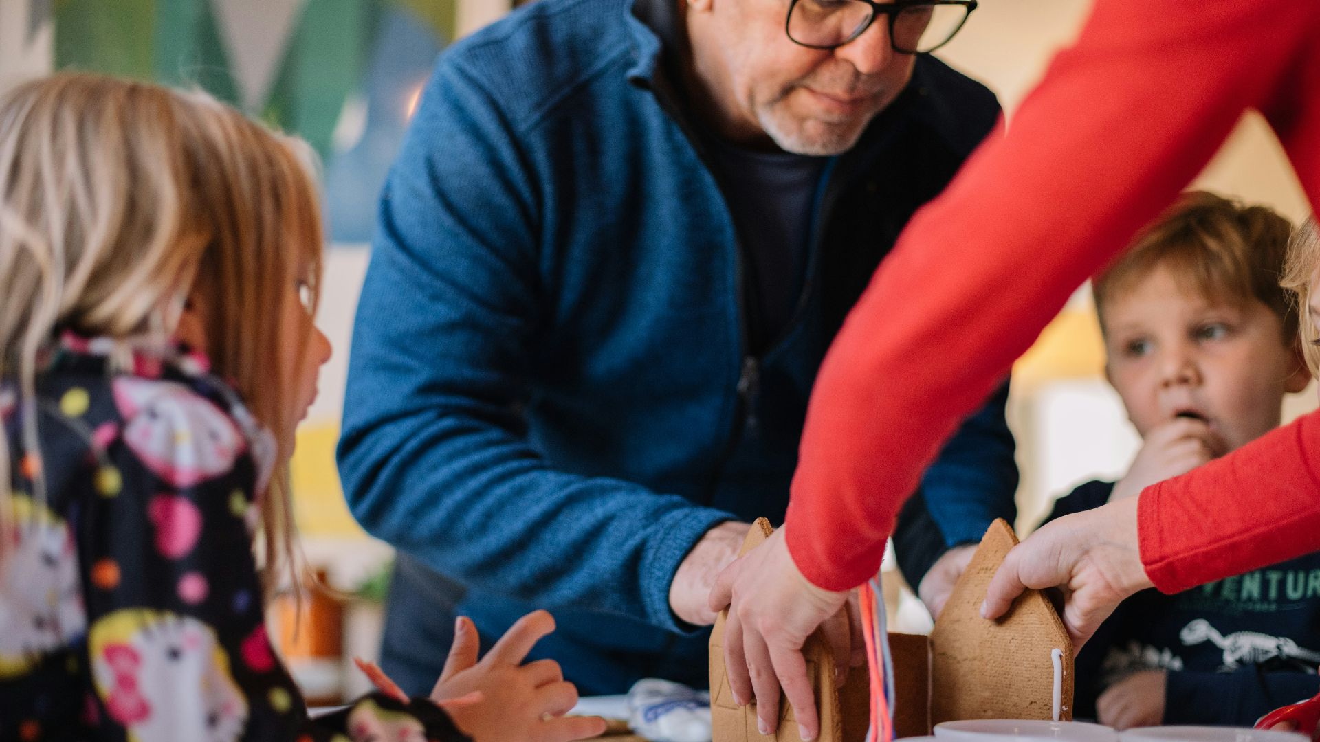 two person making gingerbread house and three children watching them