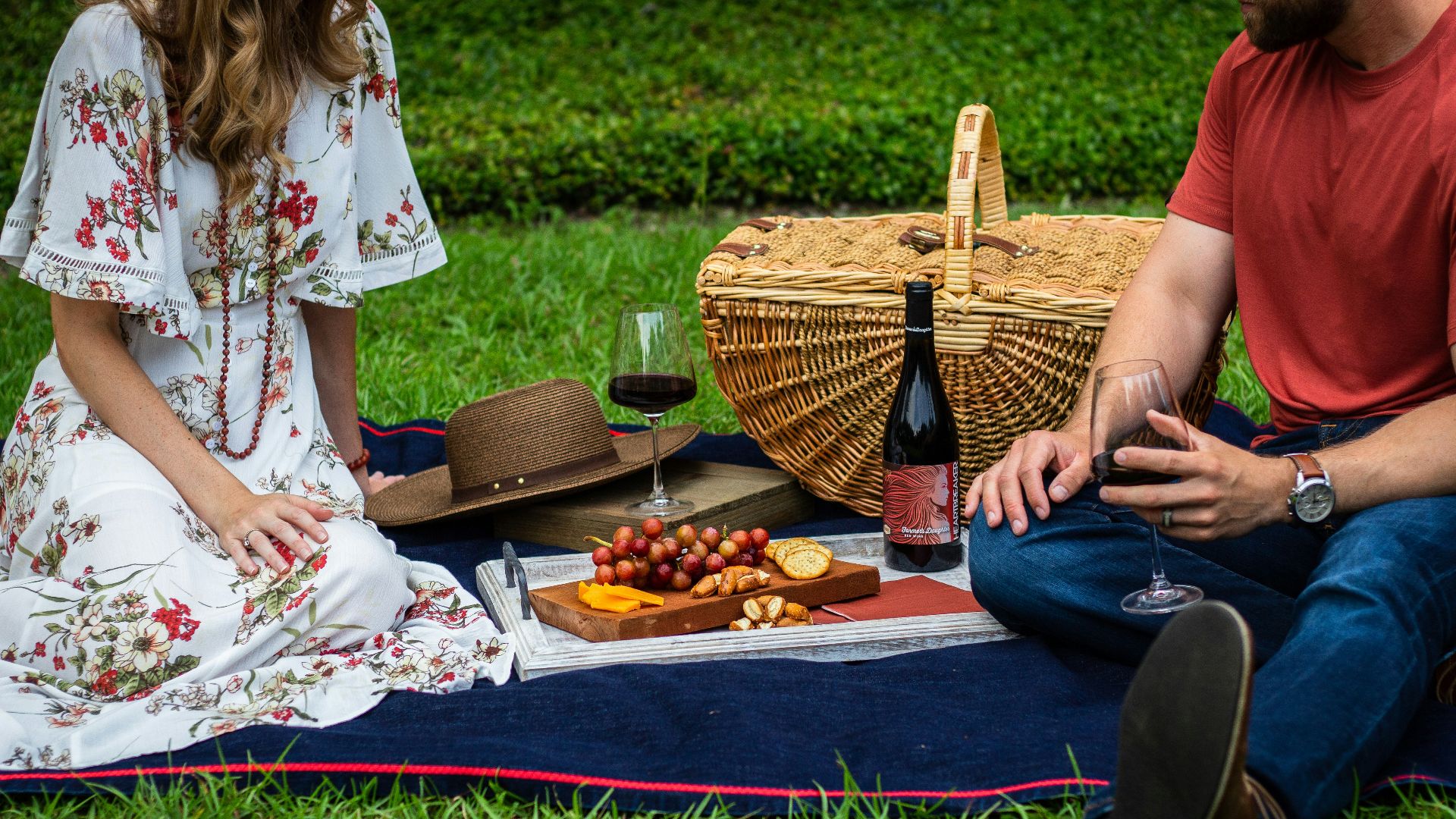 man and woman sitting on blue textile beside brown wicker picnic basket