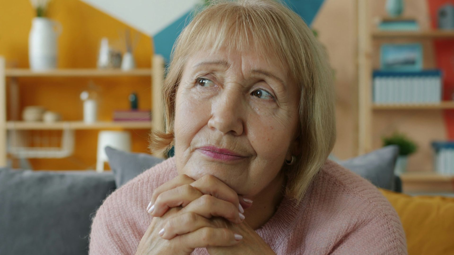 Elderly woman with hands clasped, looking thoughtful.