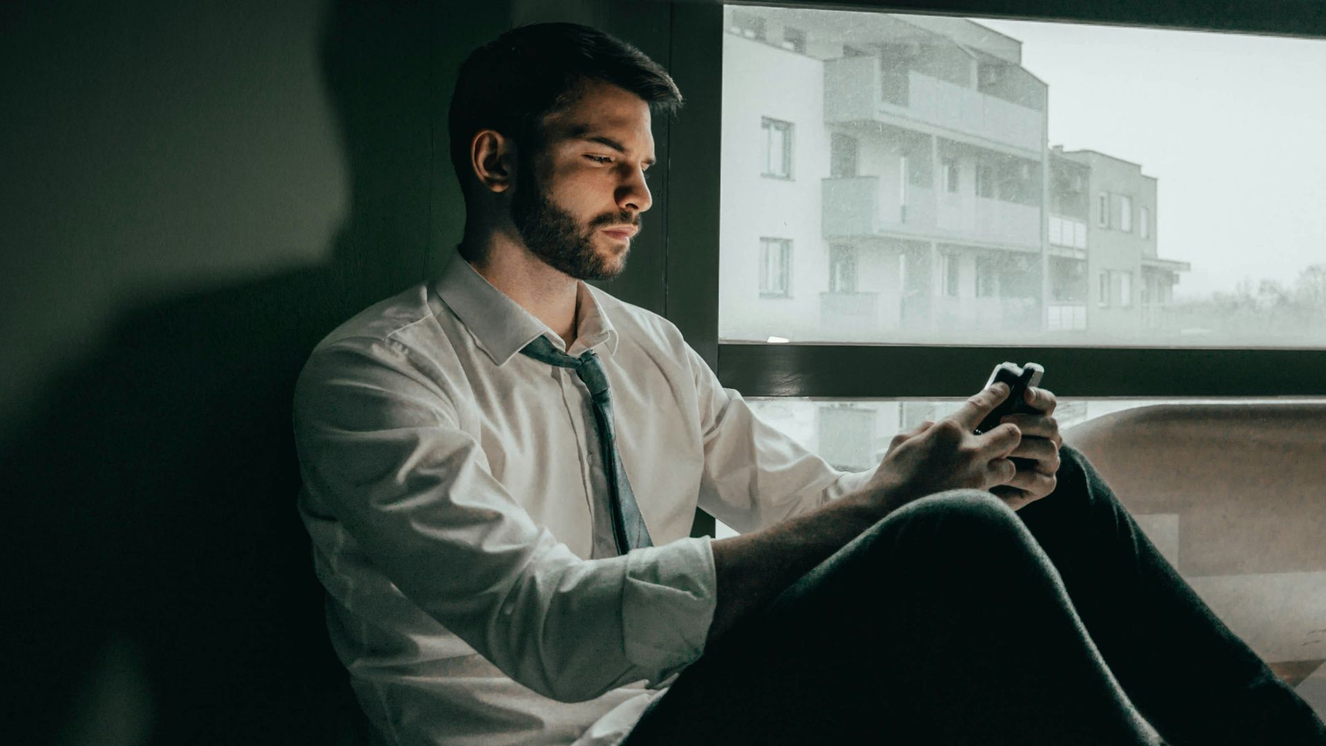 a man sitting on a window sill looking at his cell phone