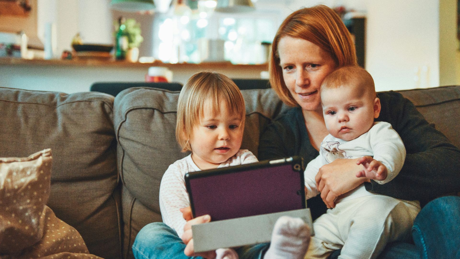two babies and woman sitting on sofa while holding baby and watching on tablet