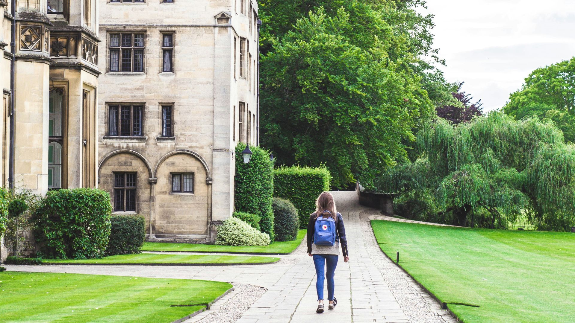 person walking on concrete road beside brown concrete house near green high trees under white sky during daytime