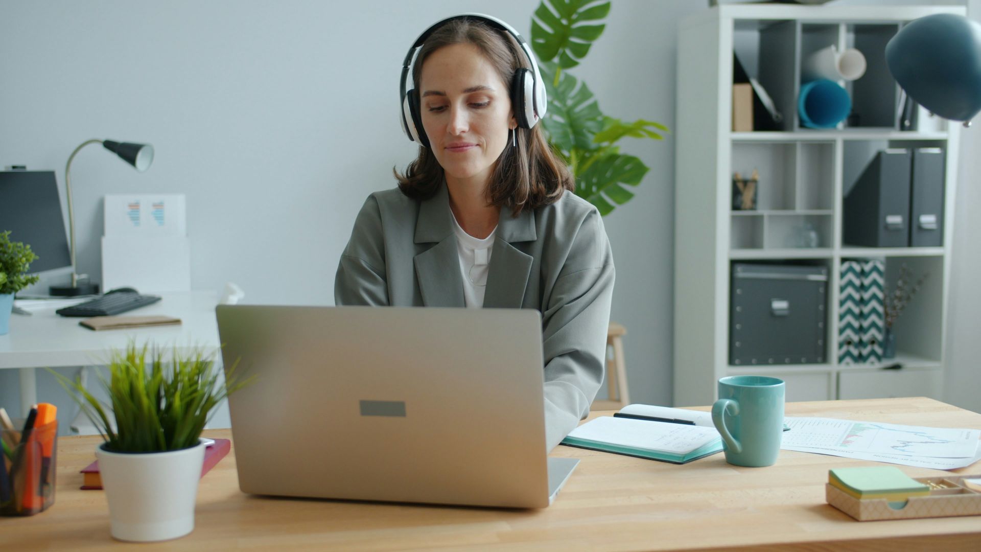 Woman wearing headphones works on laptop at desk.