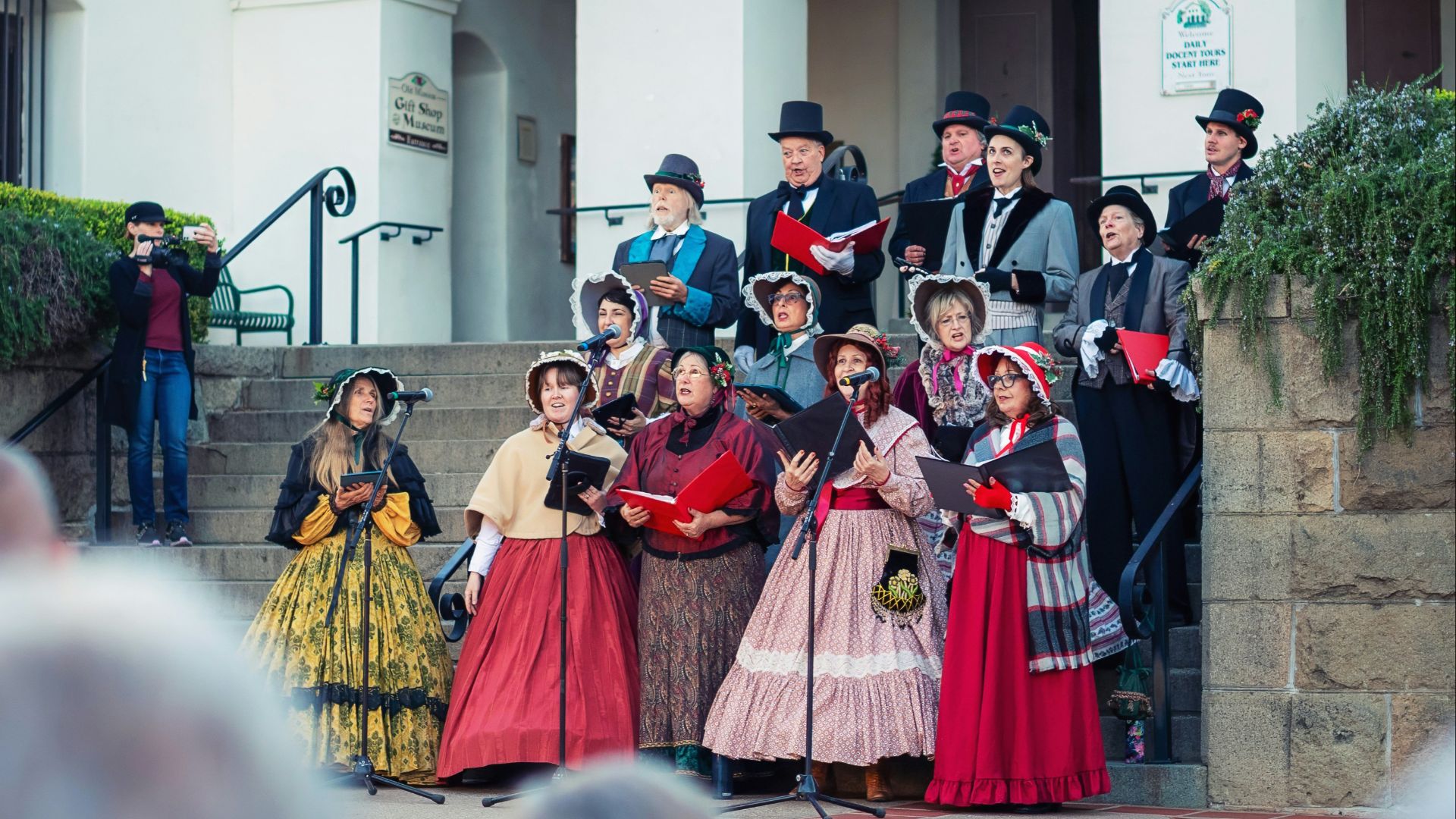 Group of carolers in vintage clothing singing on steps.