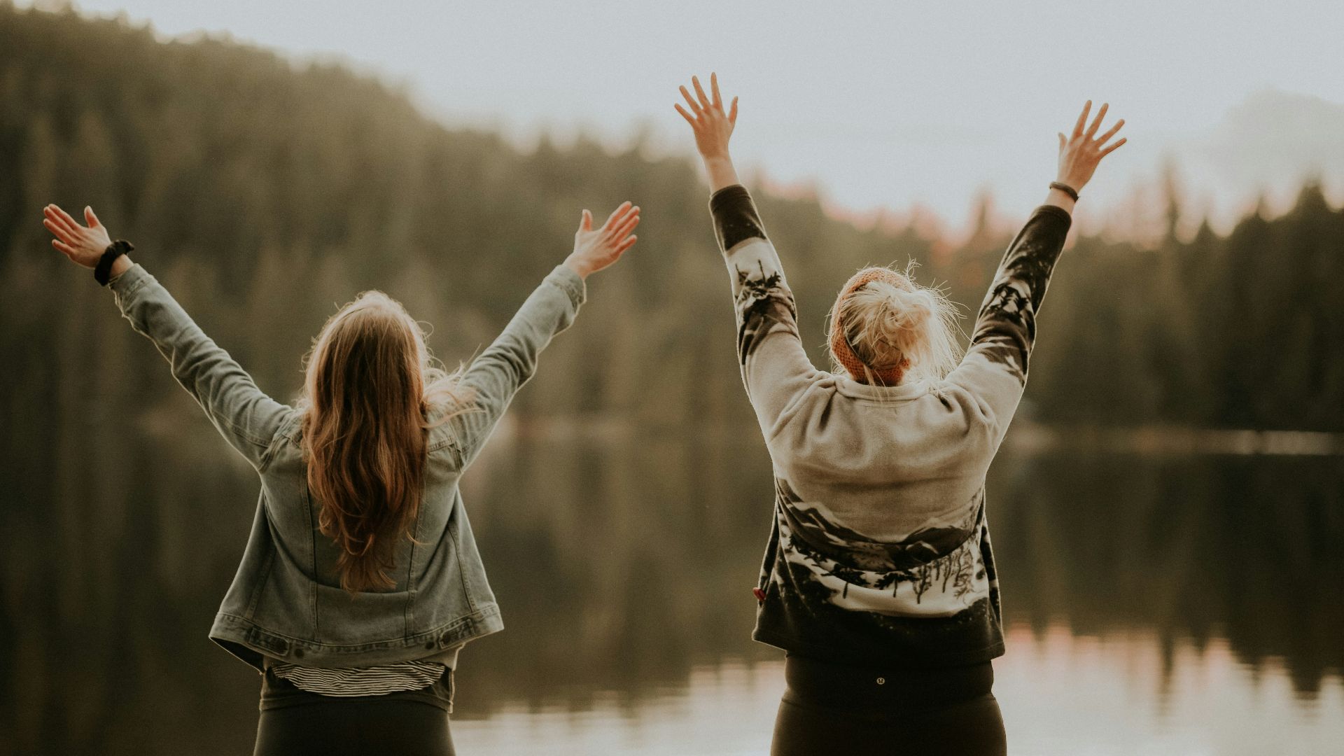 two women hands up standing beside body of water