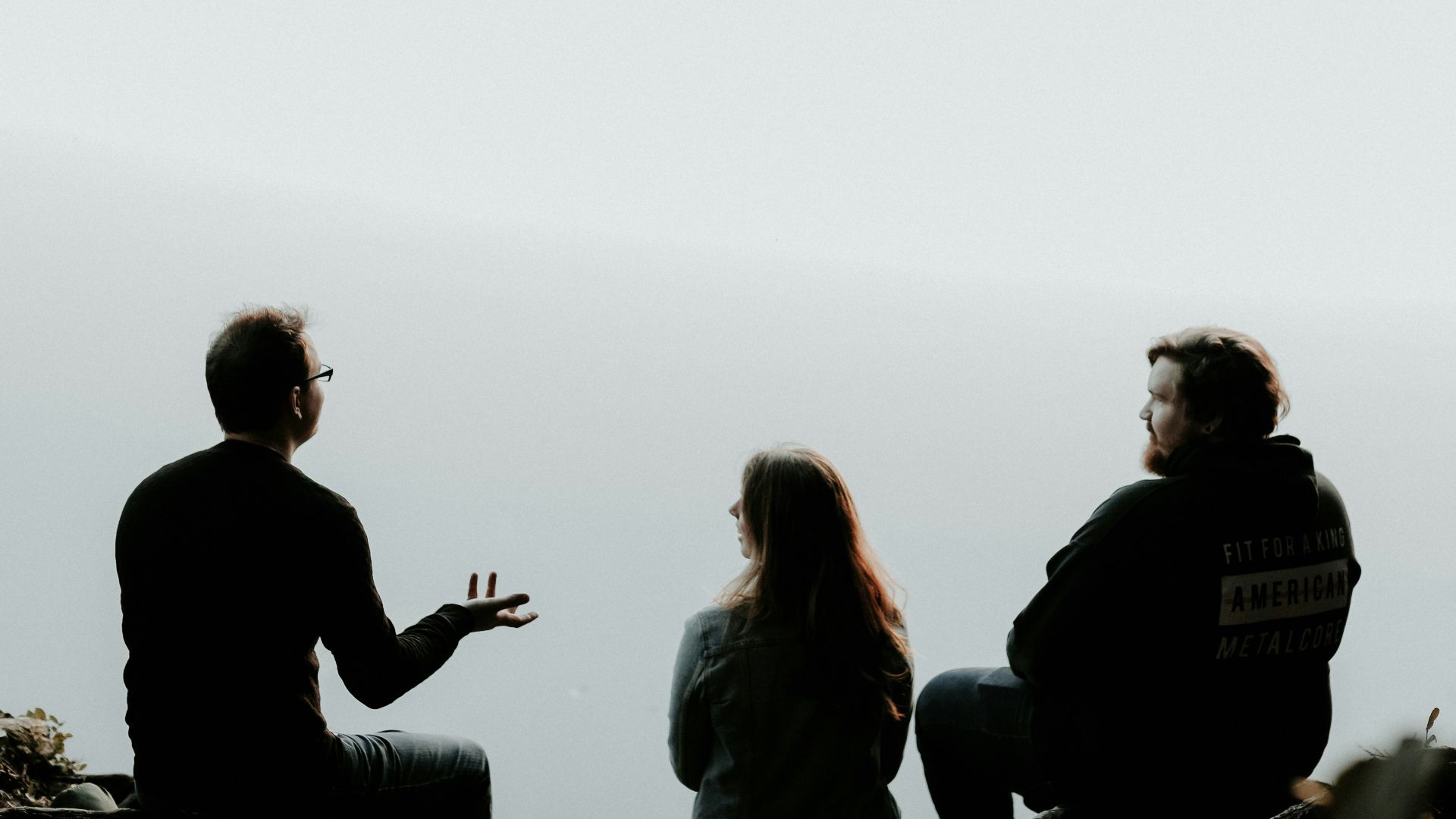 silhouette of three people sitting on cliff under foggy weather