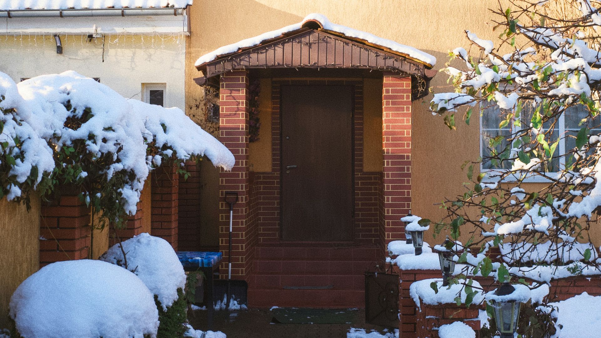 A yellow house with snow on the roof and bushes.