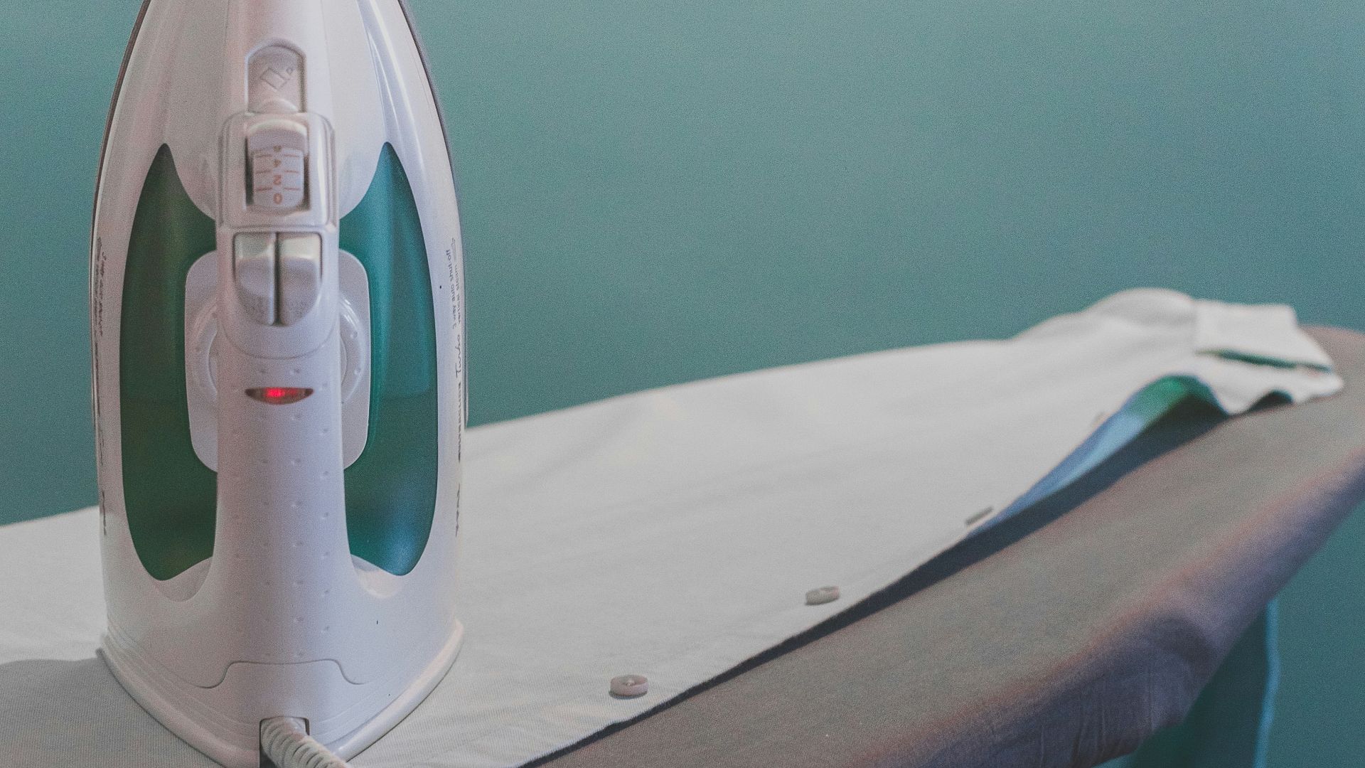 white and teal steam clothes iron plugged on ironing board