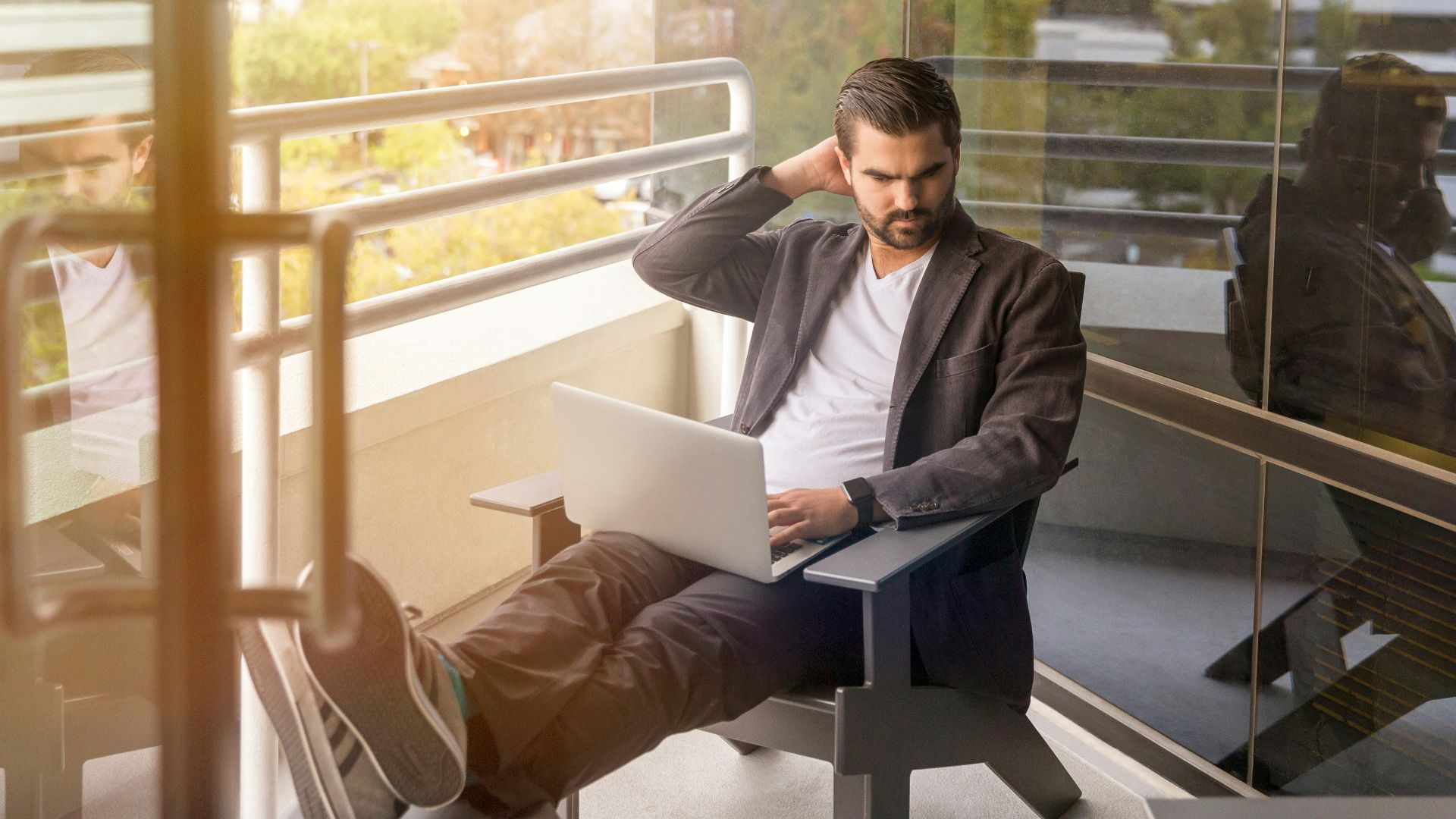 man sitting on gray arm chair using silver laptop computer on building balcony at daytime