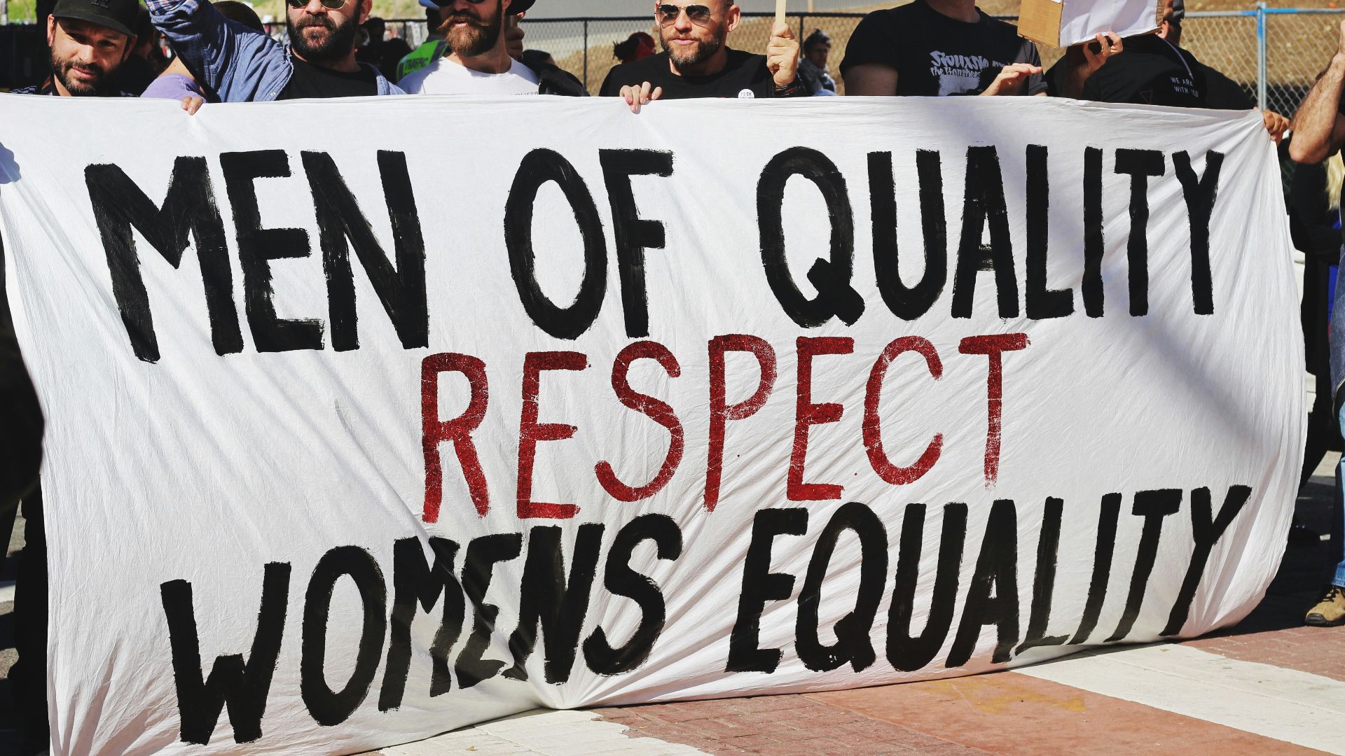 men holding white, black, and red Men of quality respect womens equality banner on road