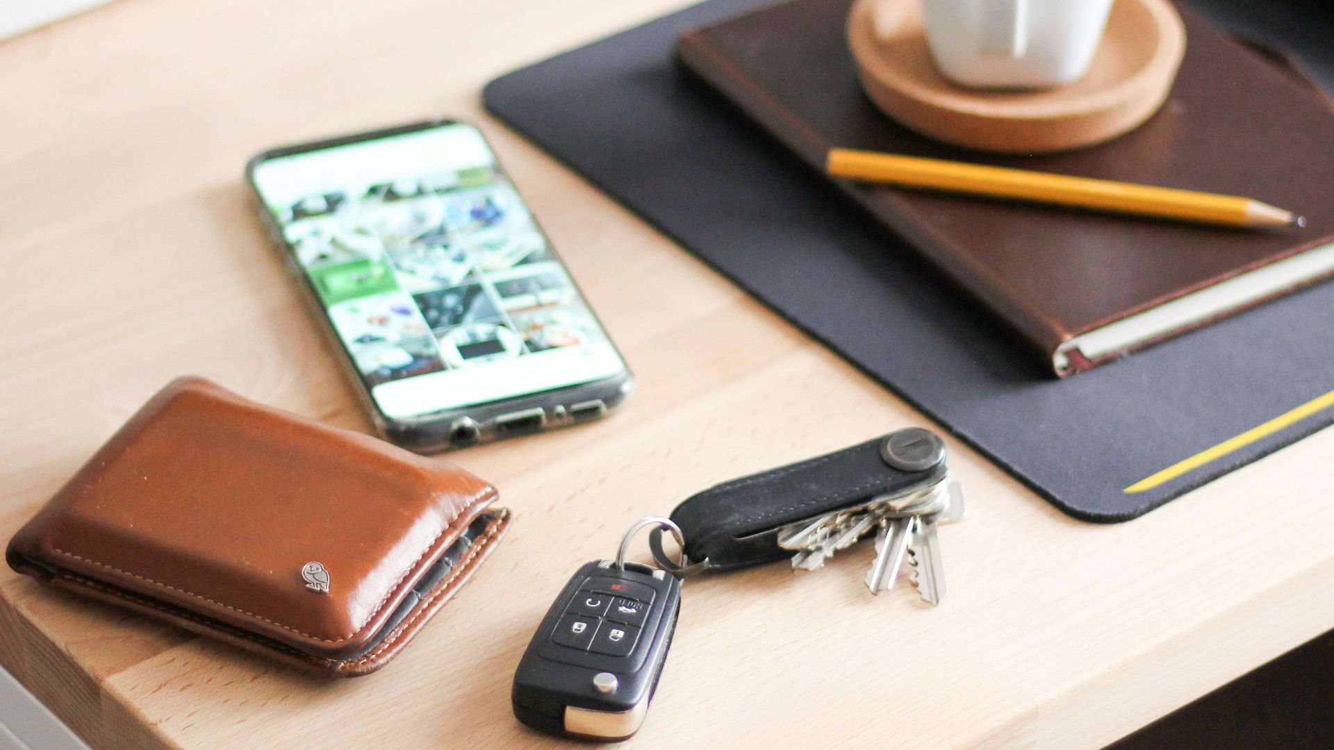 yellow wooden pencil on brown hardbound book near cappuccino, silver Android smartphone, brown leather bifold wallet, and black car fob