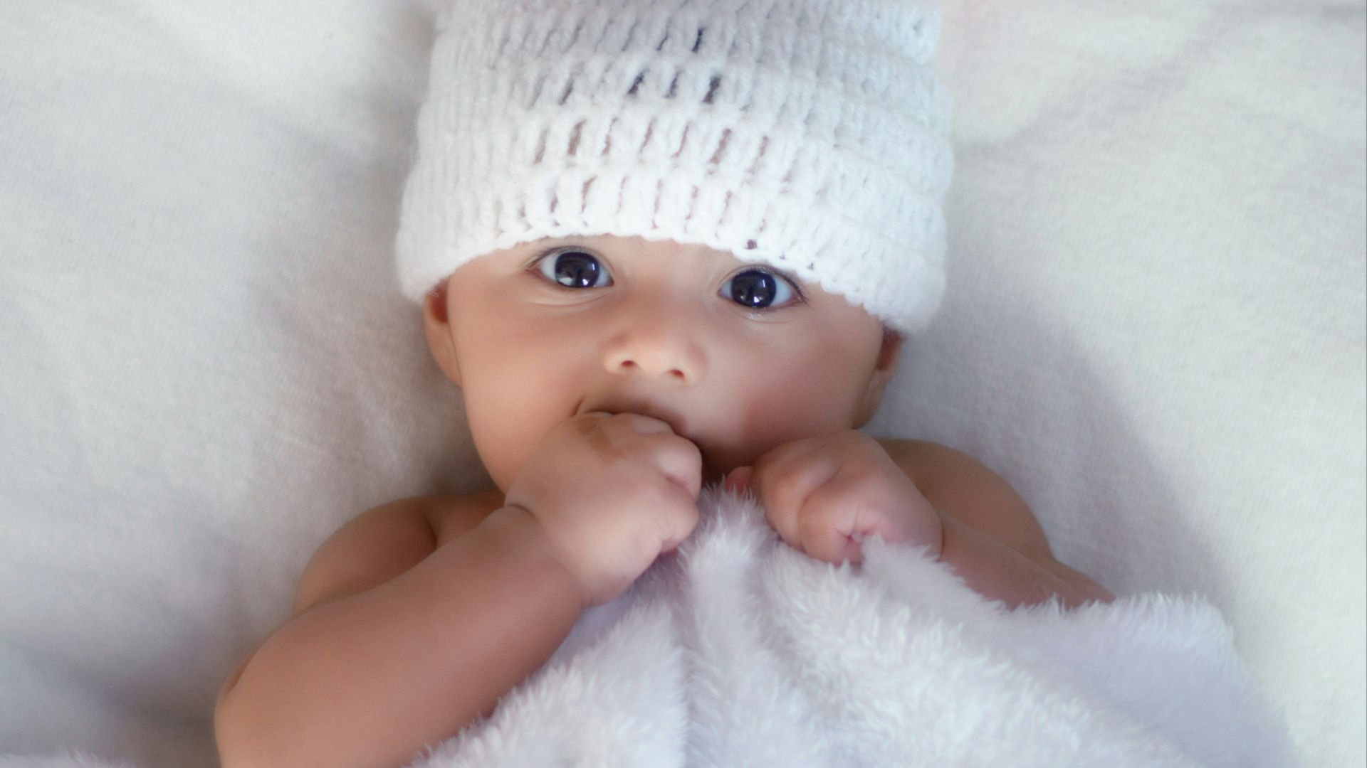 baby in white knit cap lying on white textile