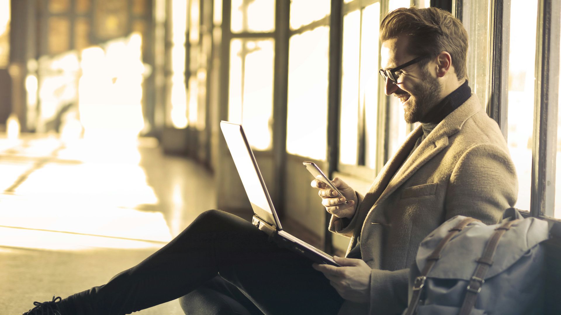 man sitting near window holding phone and laptop