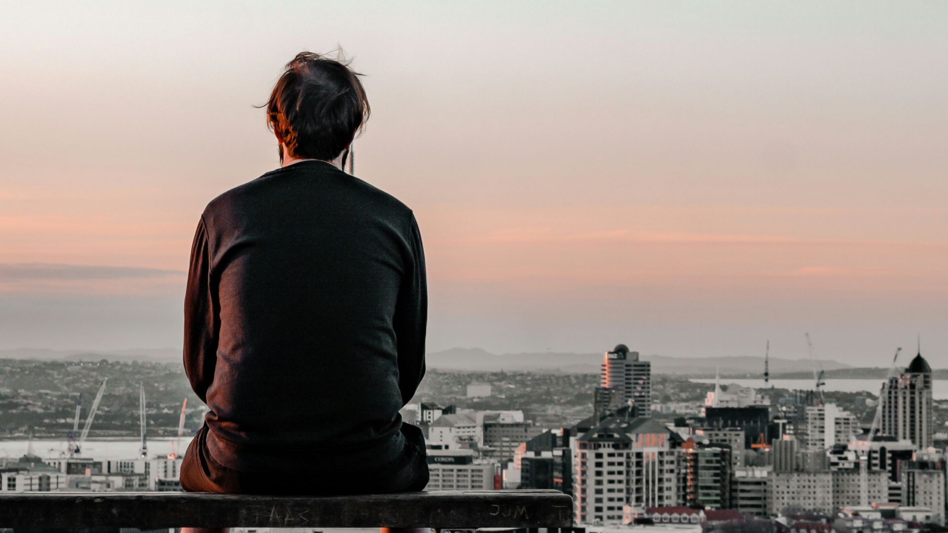 man in black long sleeve shirt sitting on brown wooden bench looking at city during daytime