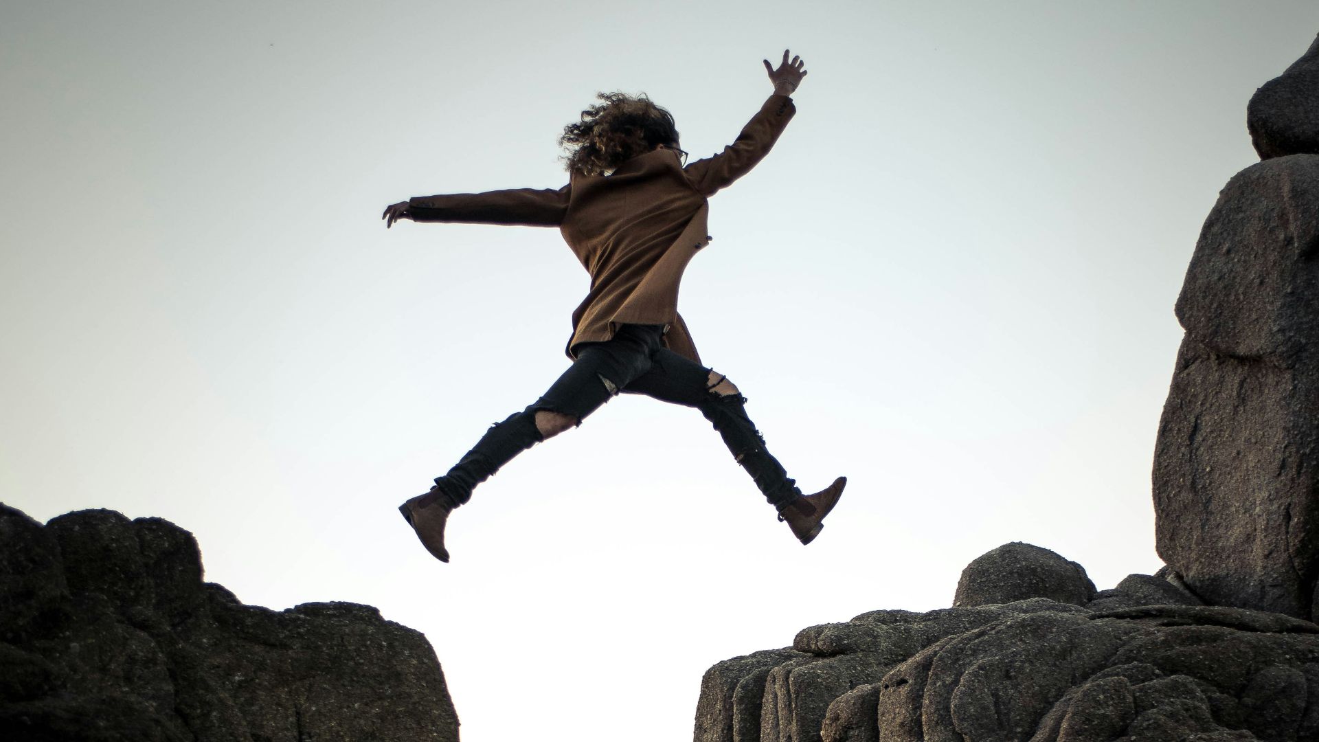 person jumping on big rock under gray and white sky during daytime