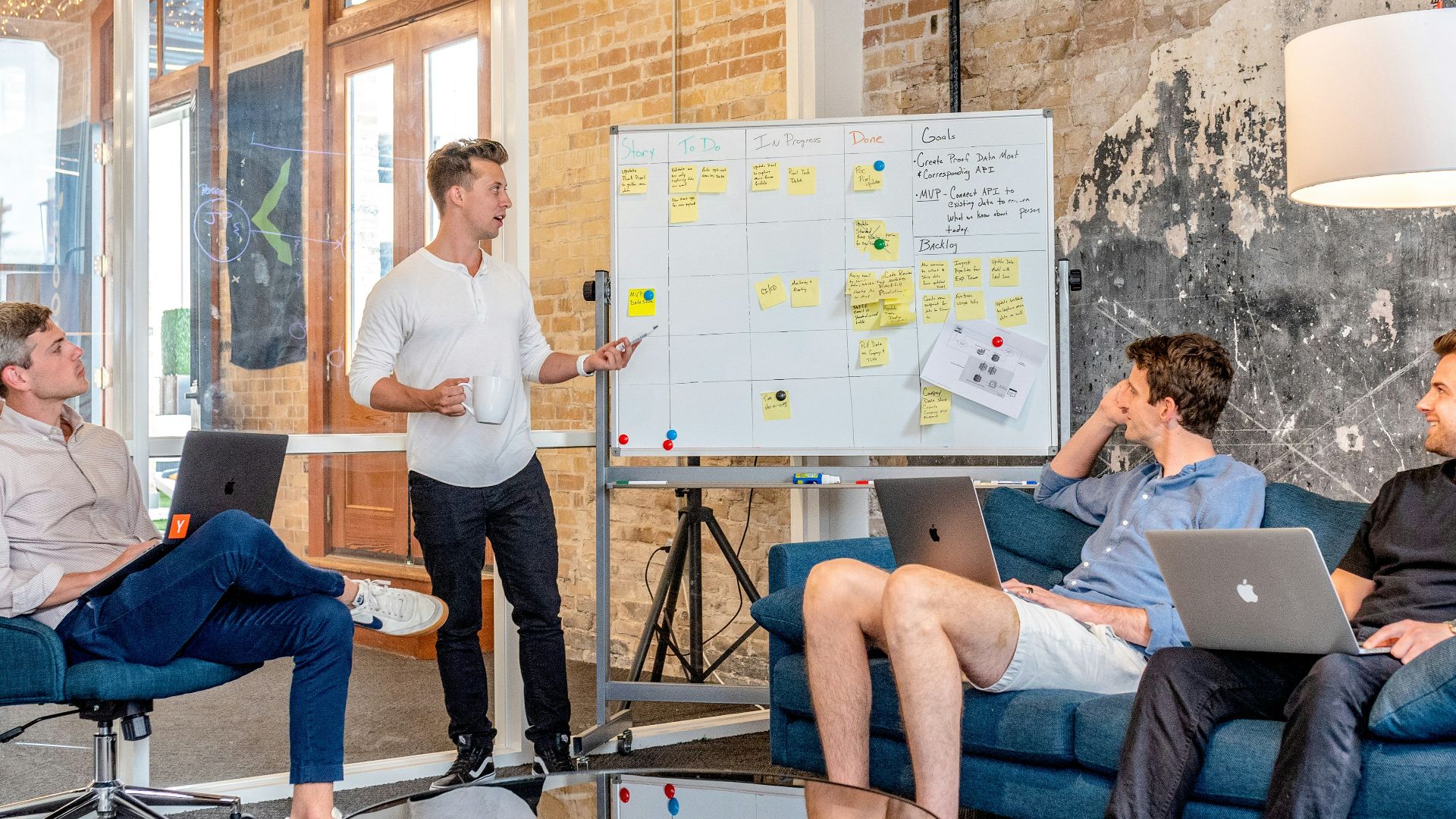 three men sitting while using laptops and watching man beside whiteboard